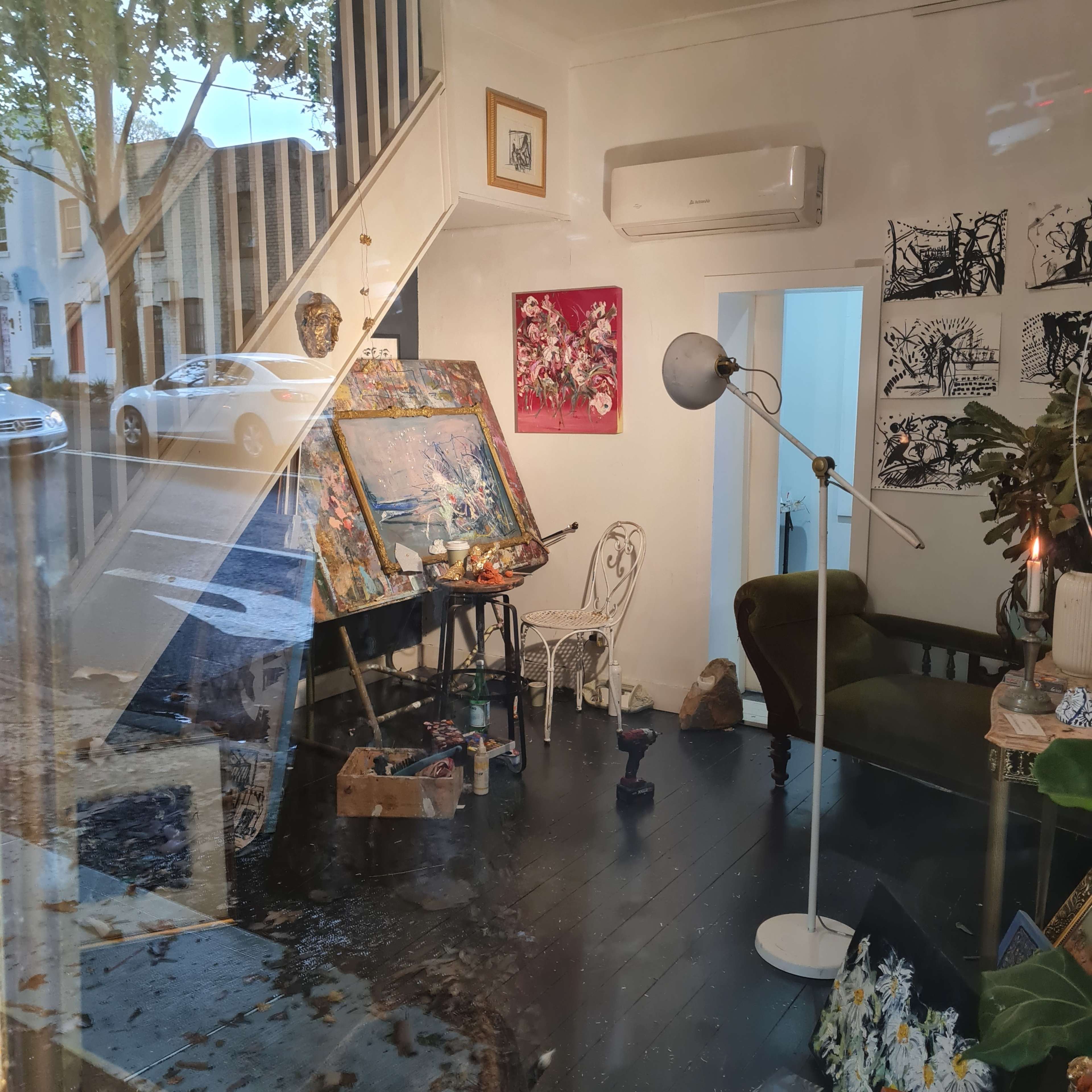 An artist's studio with an easel, a green chair, and various art materials visible through a large window, while a street scene is reflected outside.