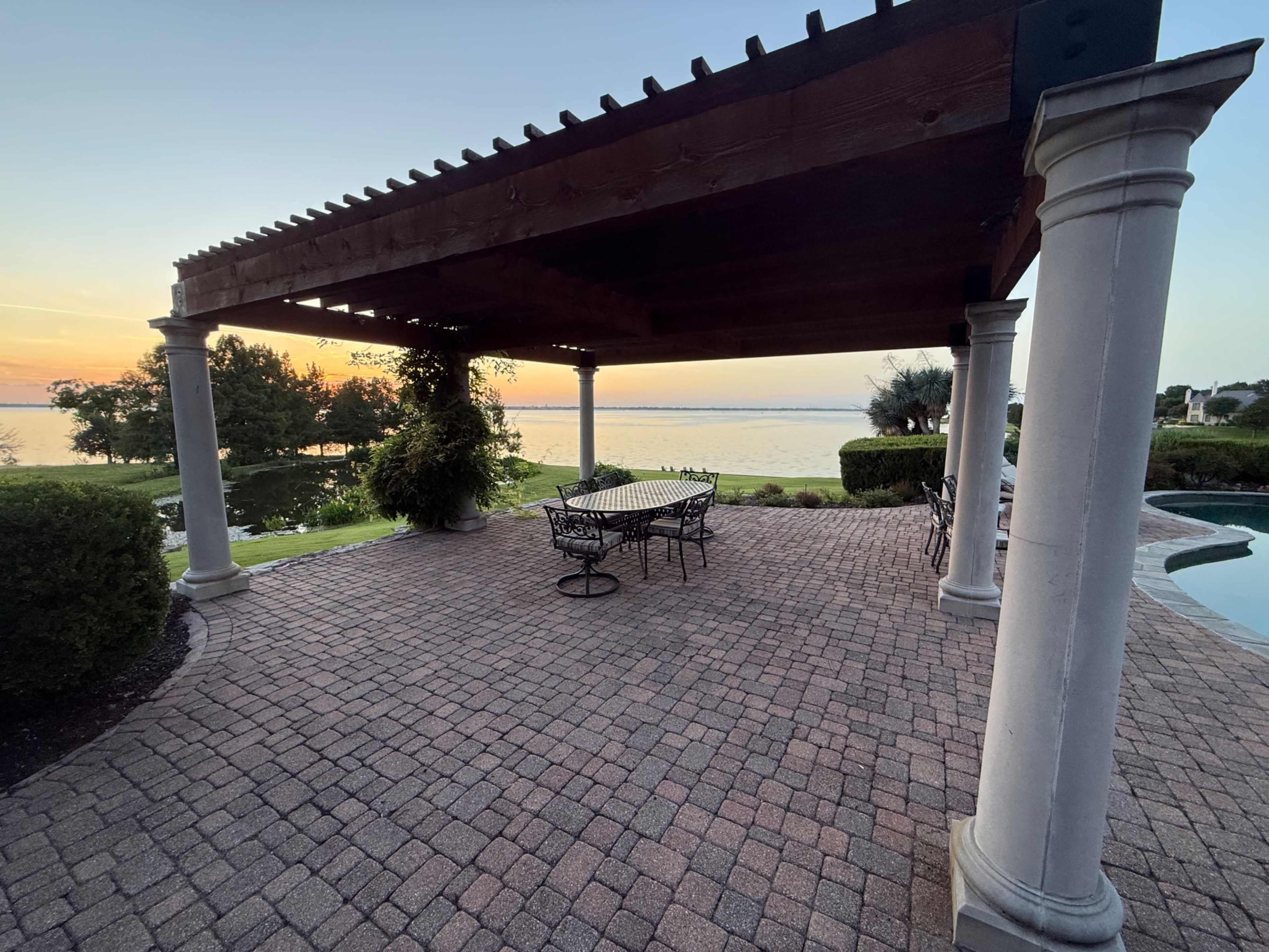A pergola with columns overlooks a patio area and a body of water at sunset.
