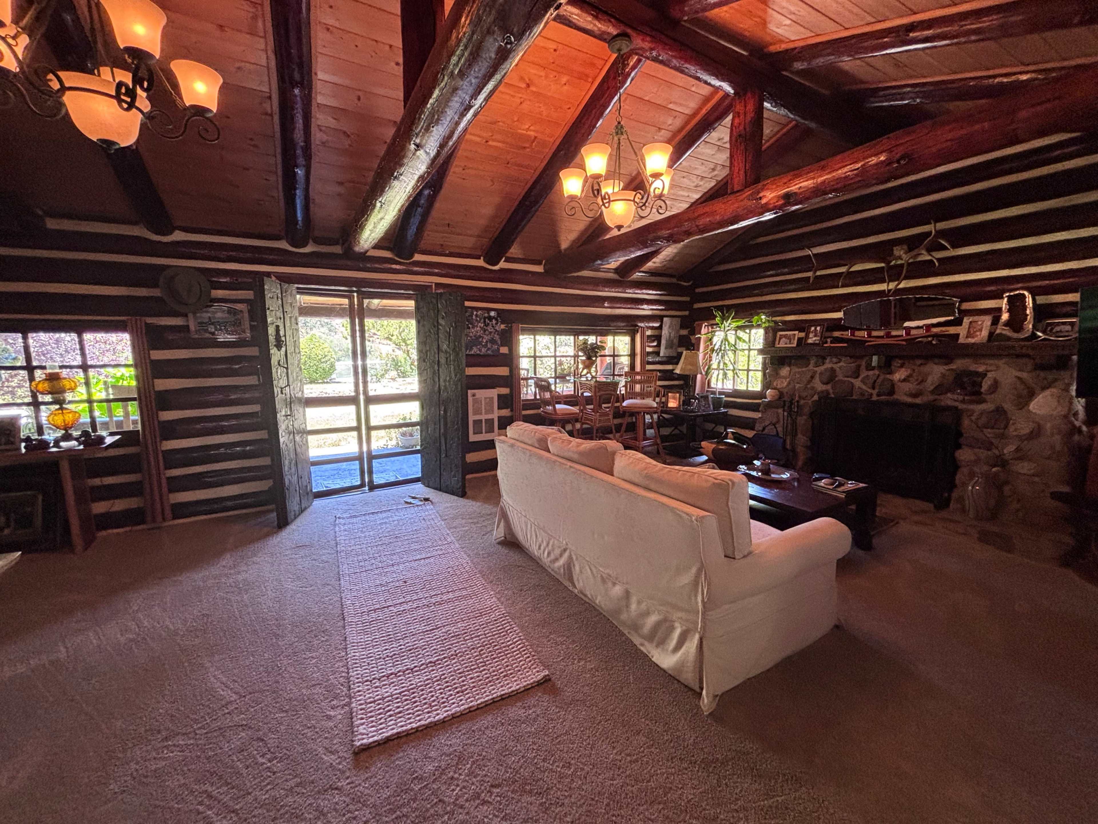 The image shows a cozy living room with wooden beams, a stone fireplace, and large windows overlooking a garden.