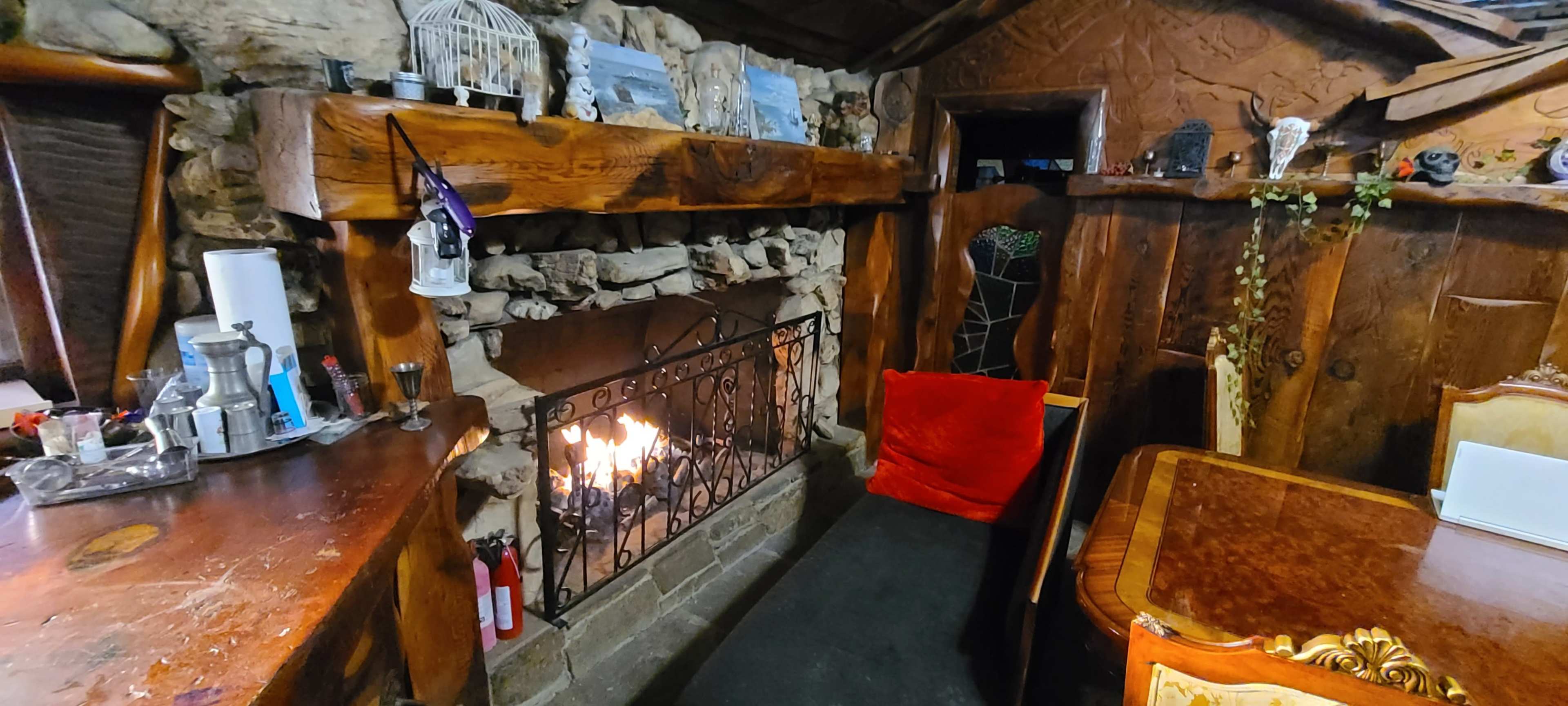 A rustic wooden fireplace with a stone mantel, featuring an open flame, is positioned near a wooden bar and a dining table in a cozy room.