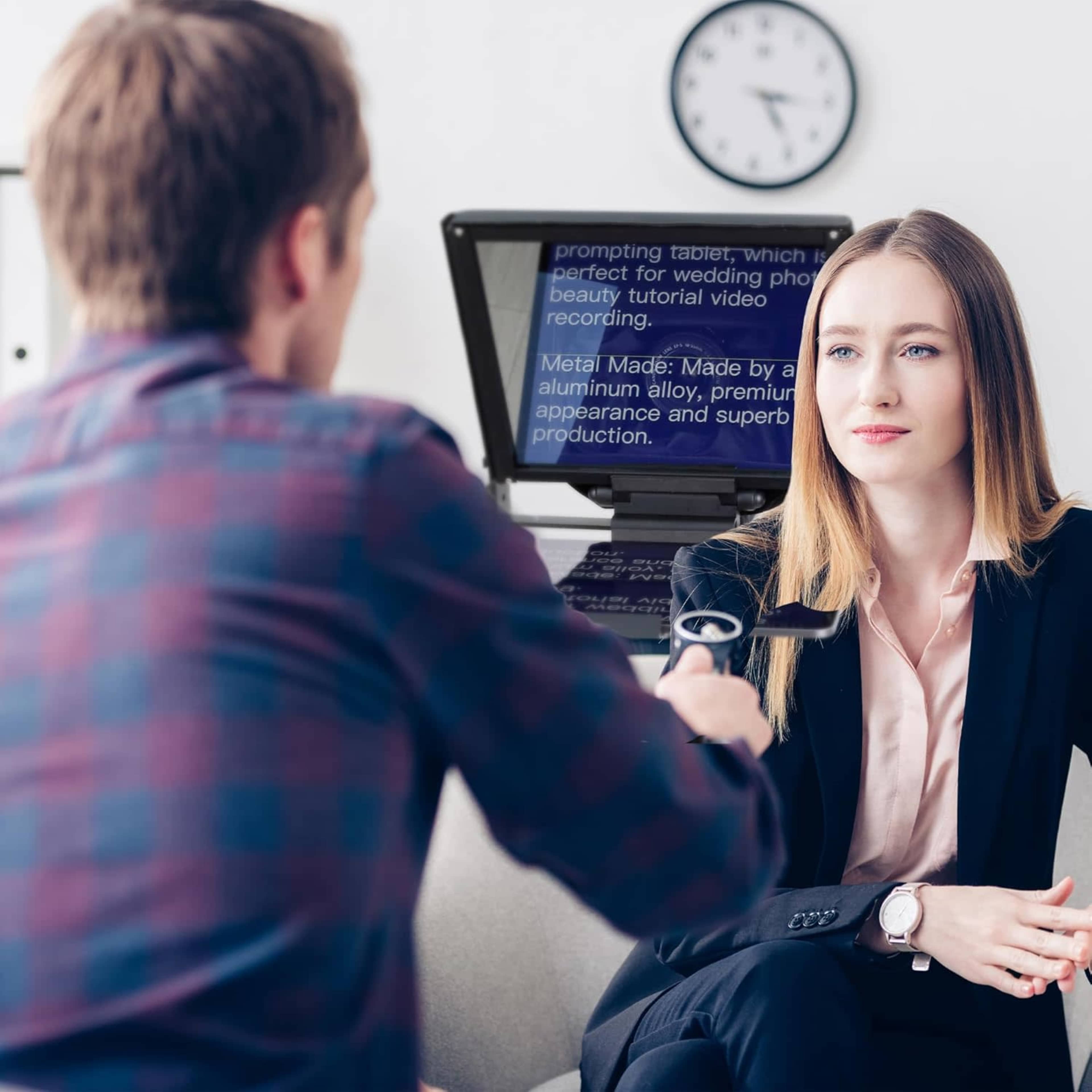 A woman in a business suit sits across from a man who is holding a microphone, with a tablet displaying text in the background.
