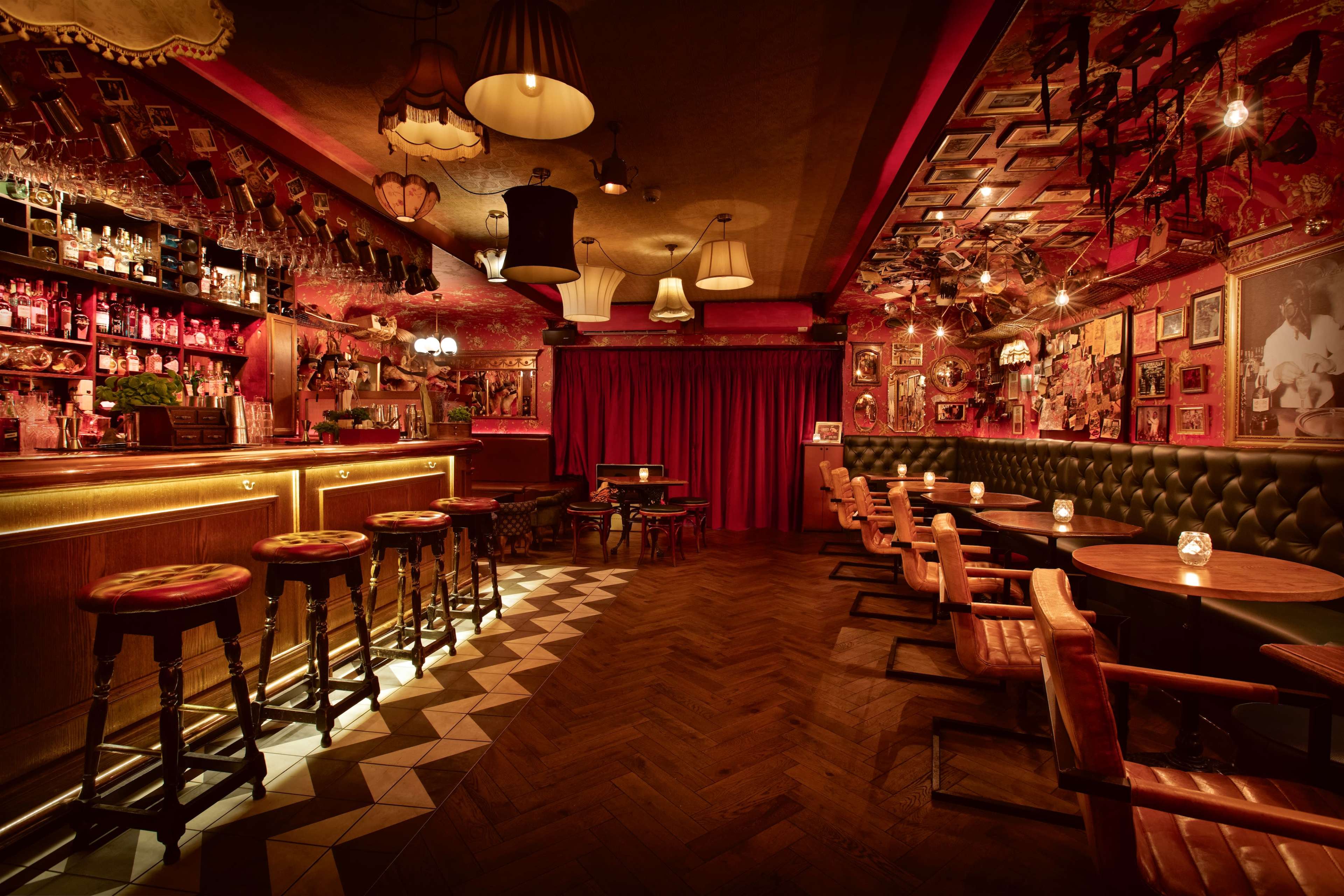 The image shows a dimly lit bar with a wooden counter, high stools, and a variety of framed pictures on the red walls.