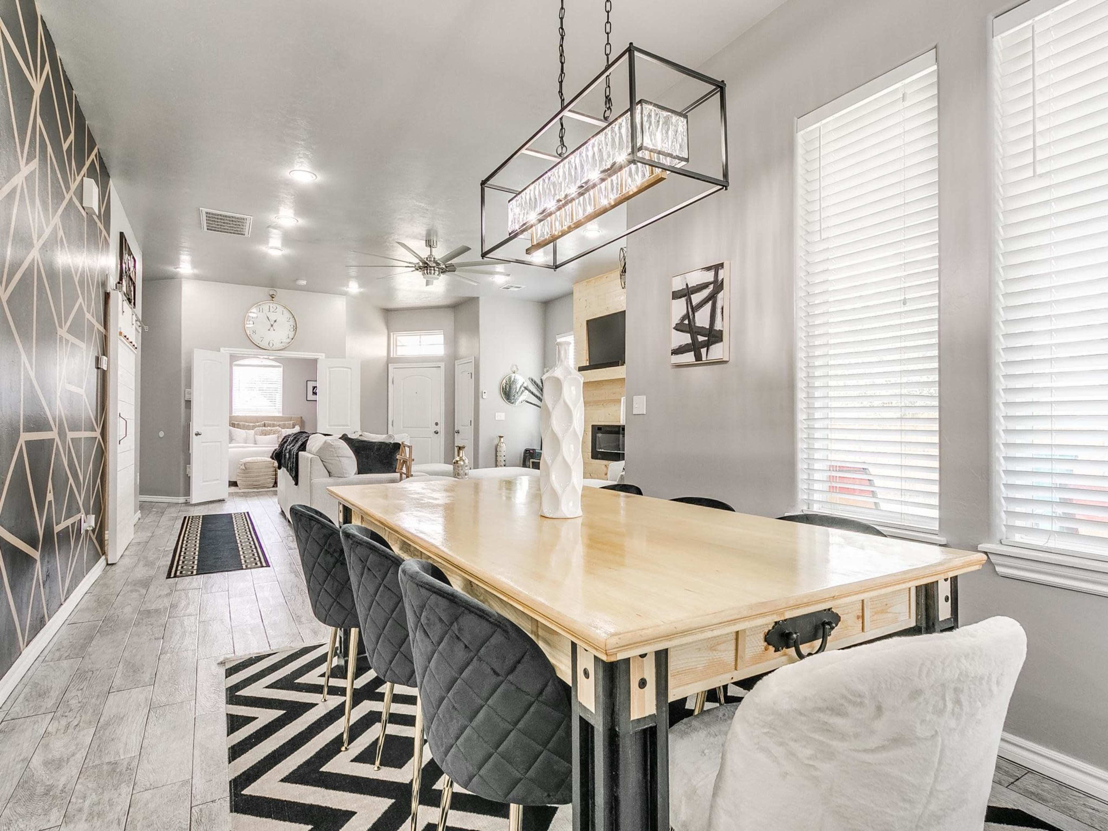 A modern dining area features a large wooden table with black chairs, surrounded by light gray walls and large windows, leading into an open living space.