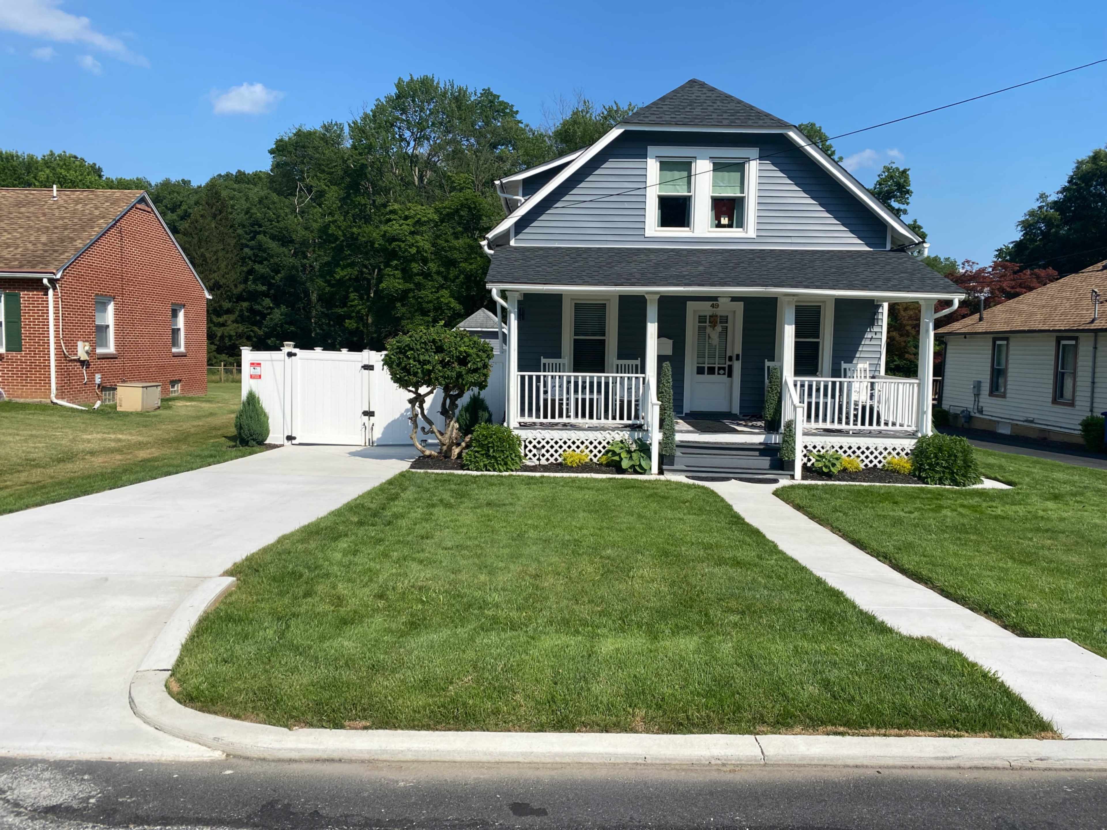 A light blue two-story house with a front porch and a driveway lined by a manicured lawn and shrubs is situated on a residential street.