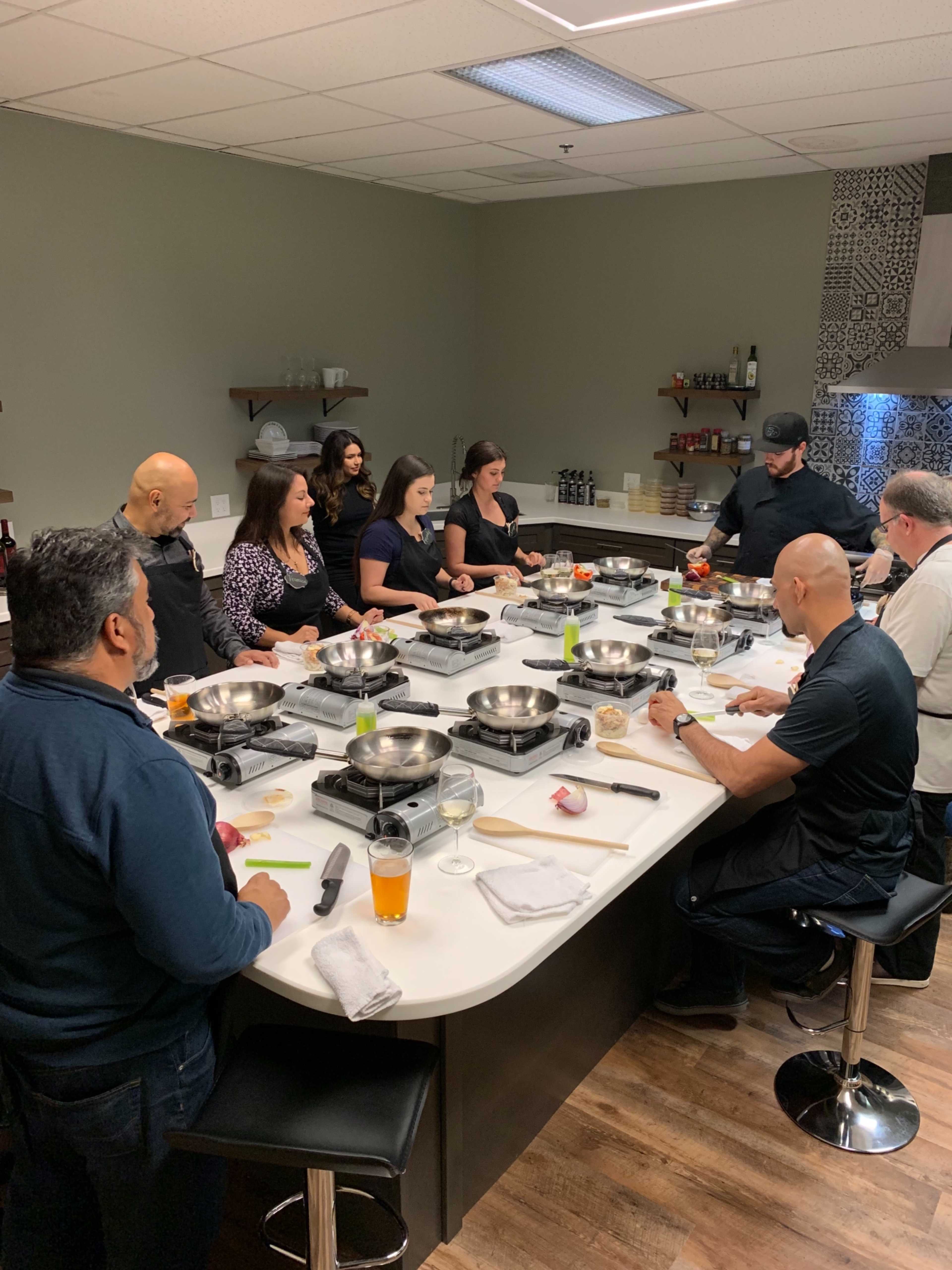 A group of people is gathered around a kitchen island, preparing food using various cooking tools and ingredients.