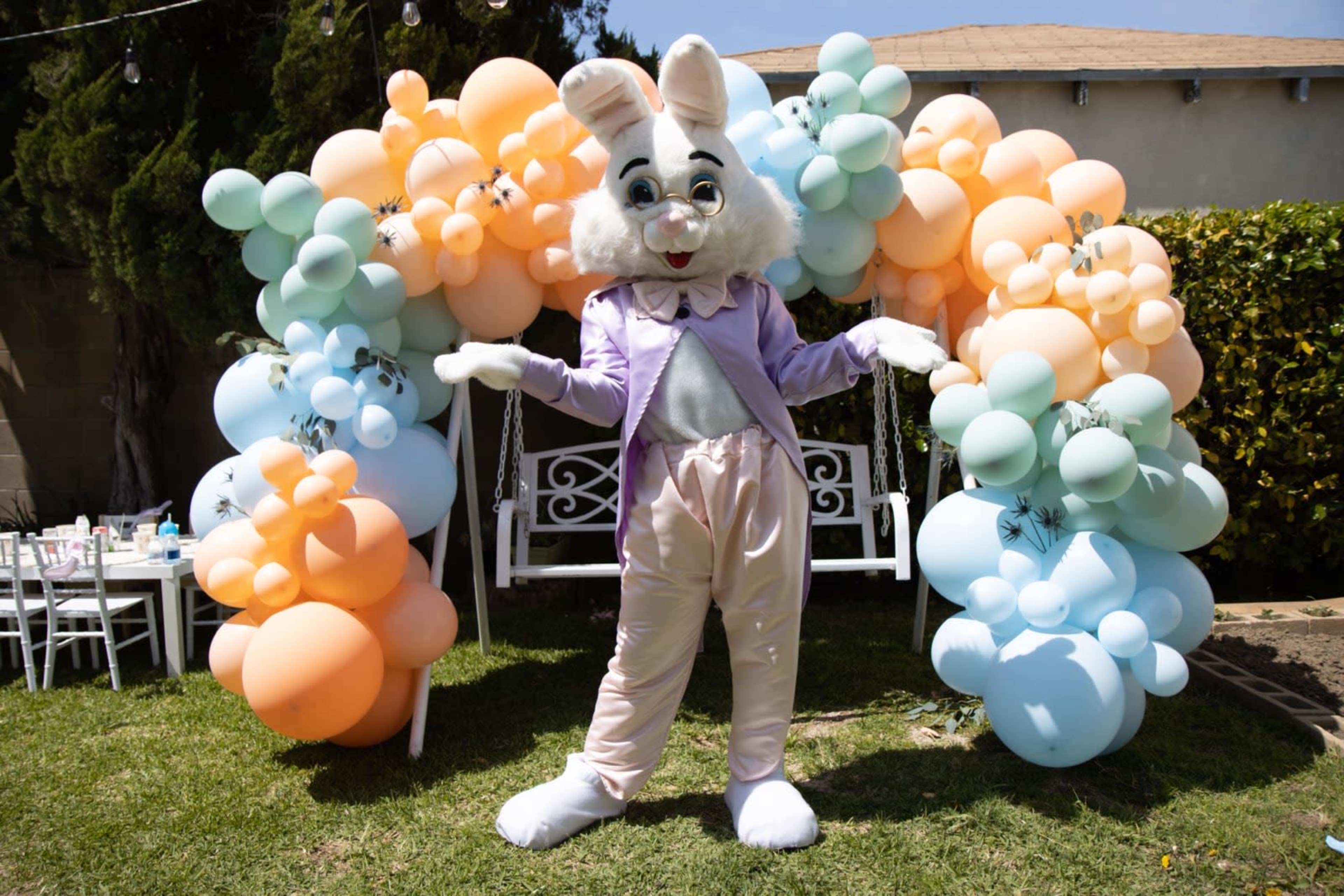 A person in a bunny costume stands in front of a colorful balloon arch at an outdoor event.