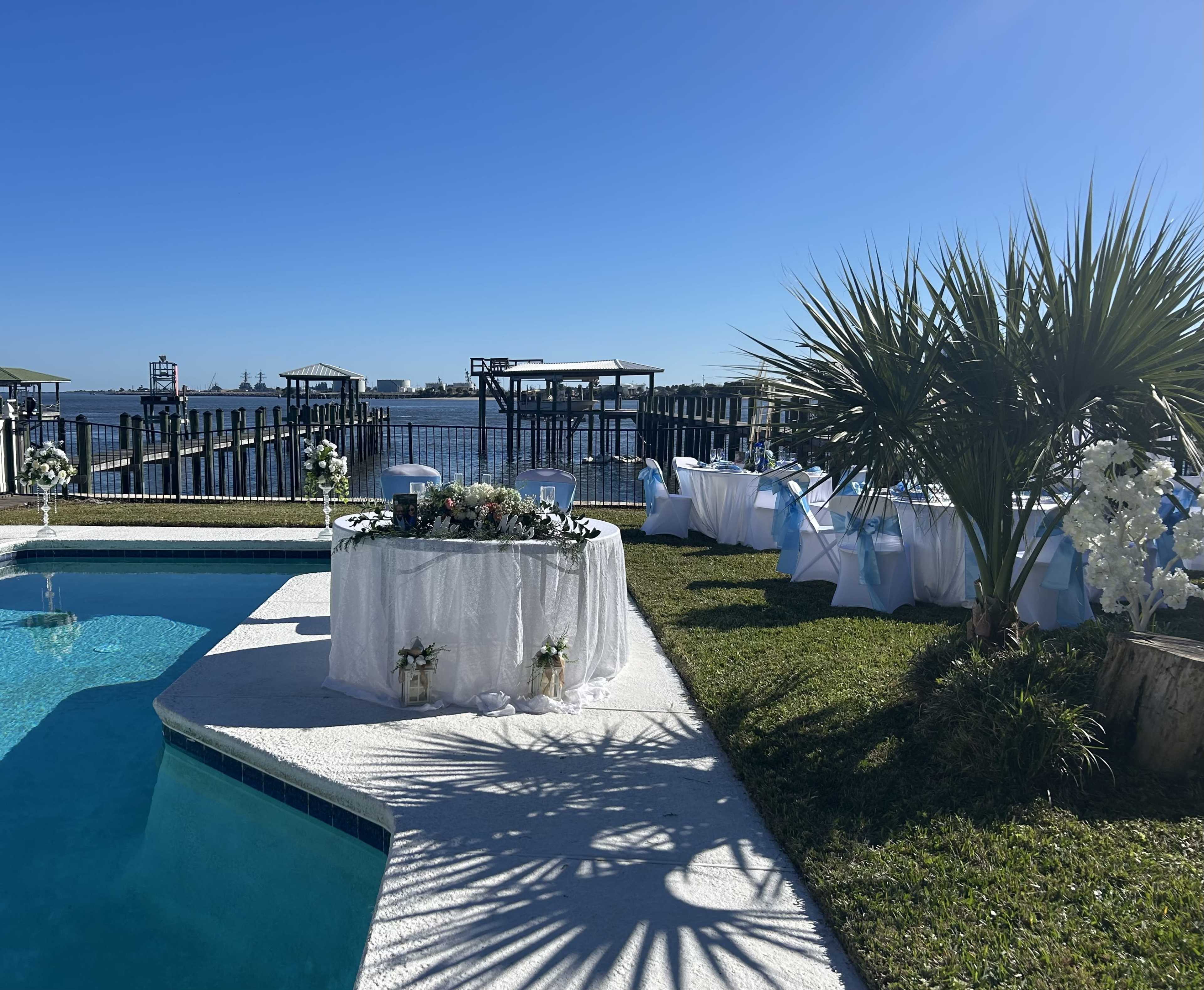 A decorated table with floral arrangements is set beside a pool overlooking a waterfront with nearby docks.
