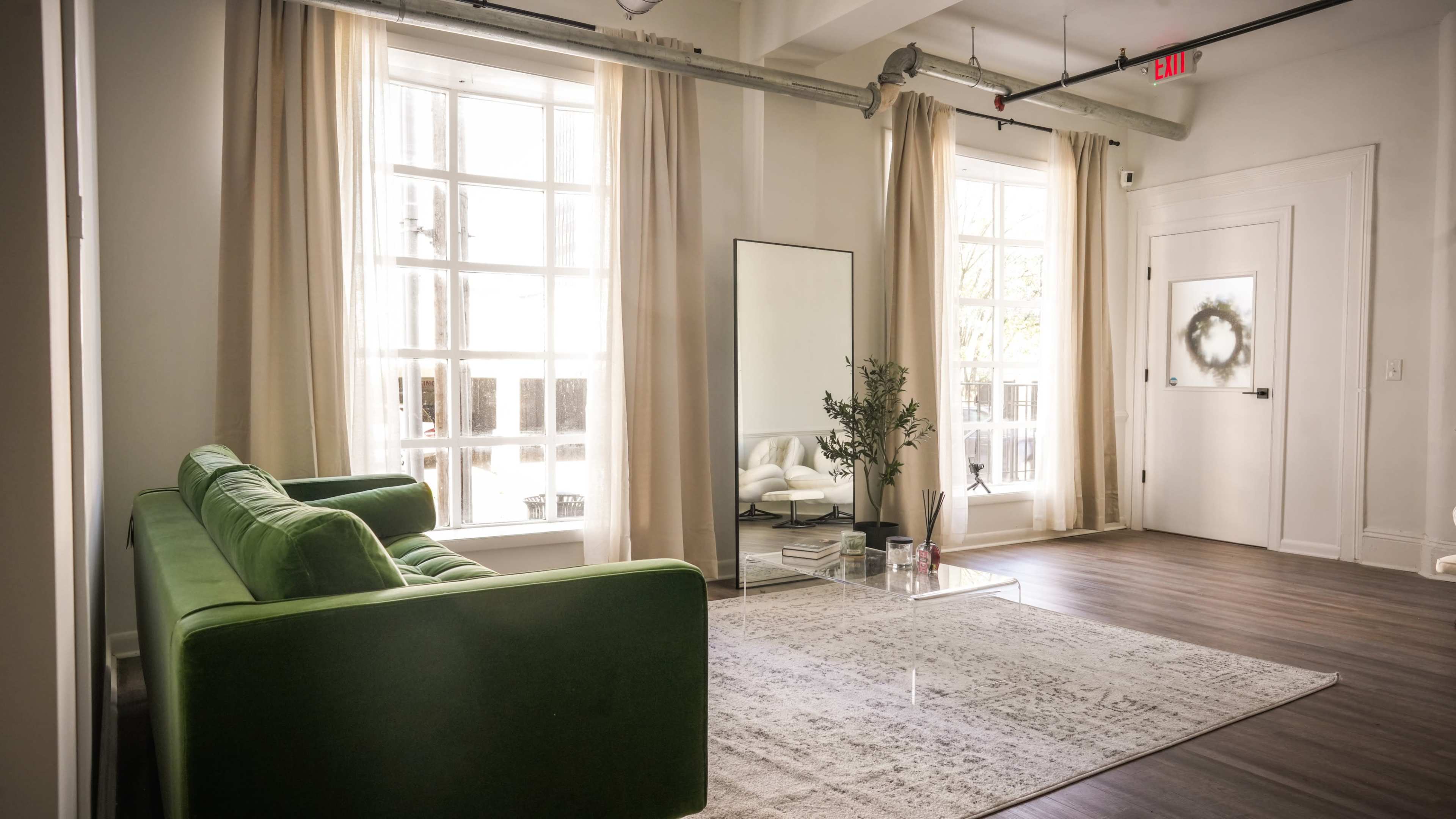 A green sofa is positioned across from a mirror and a small table on a light-colored area rug in a well-lit living space with large windows and pale curtains.