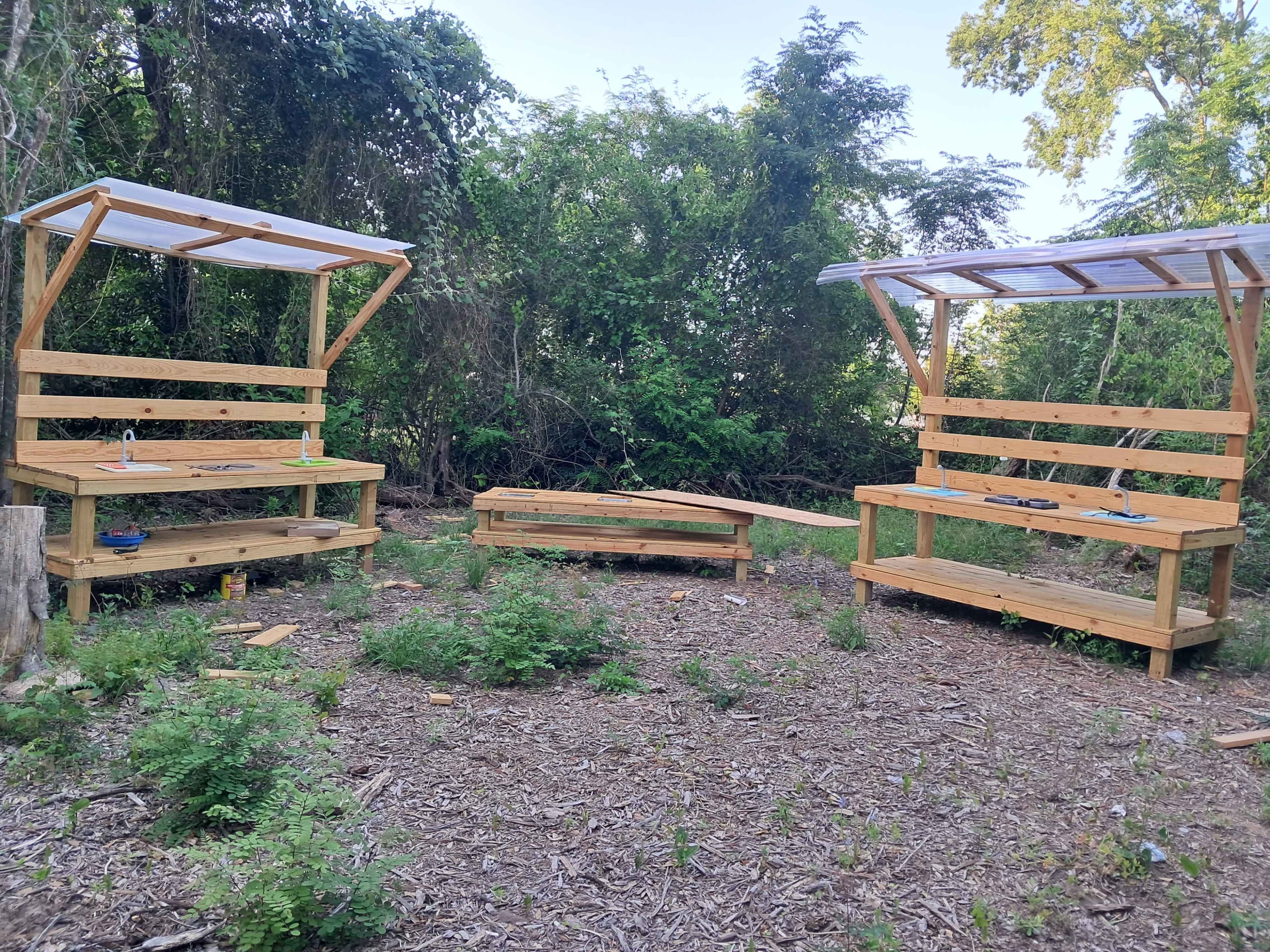 The image shows two wooden picnic-style benches with roofs, placed in a clearing surrounded by dense greenery.
