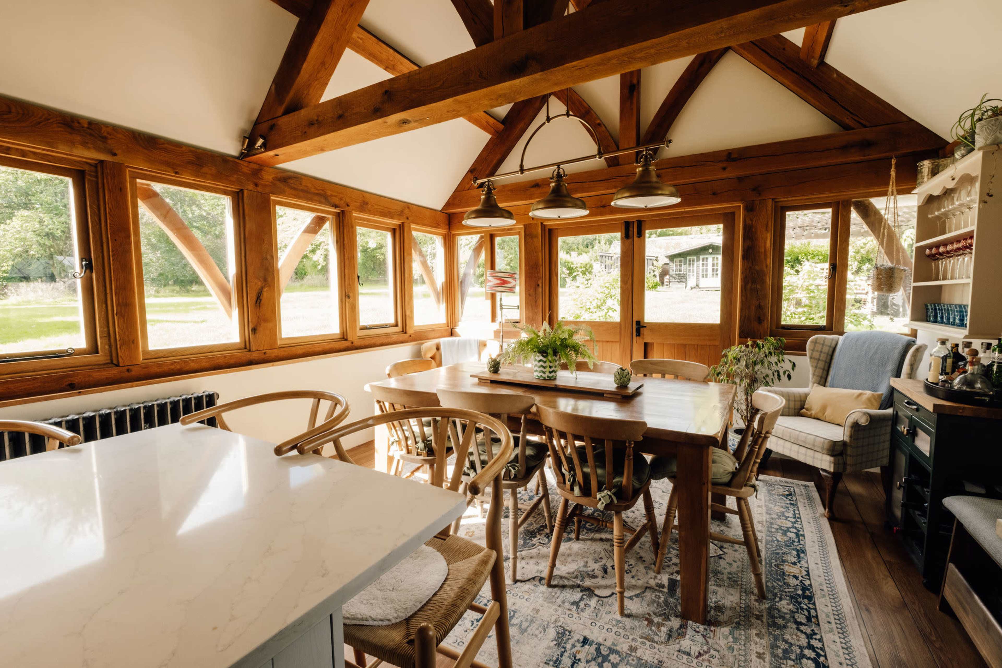 The image shows a spacious dining area with wooden beams, large windows providing natural light, and a long wooden table surrounded by chairs.