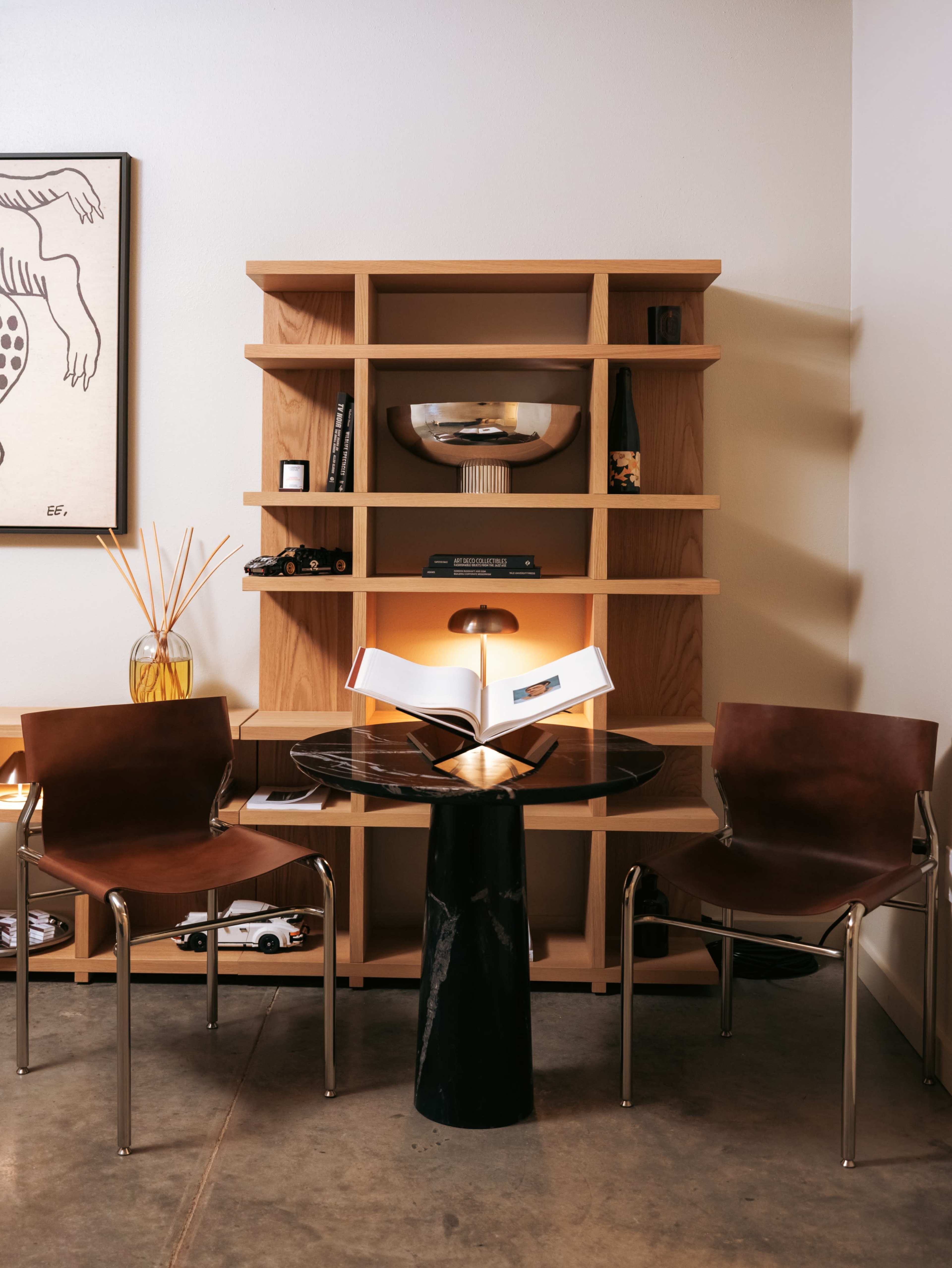 The image shows a minimalistic interior with a wooden shelf, a round black table, two brown chairs, and an open book positioned in front of a warm light.