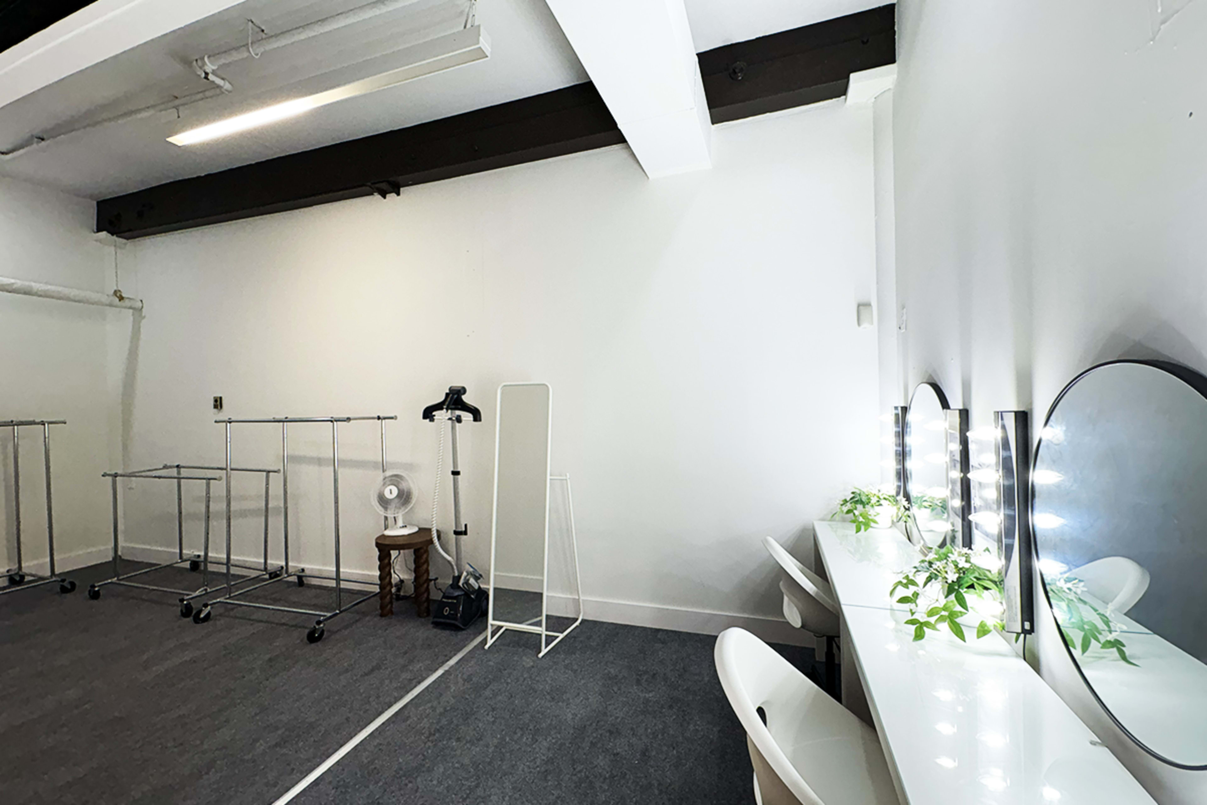 The image shows a minimalist dressing room with mirrors, a fan, a tall mirror, and clothing racks against a white wall.