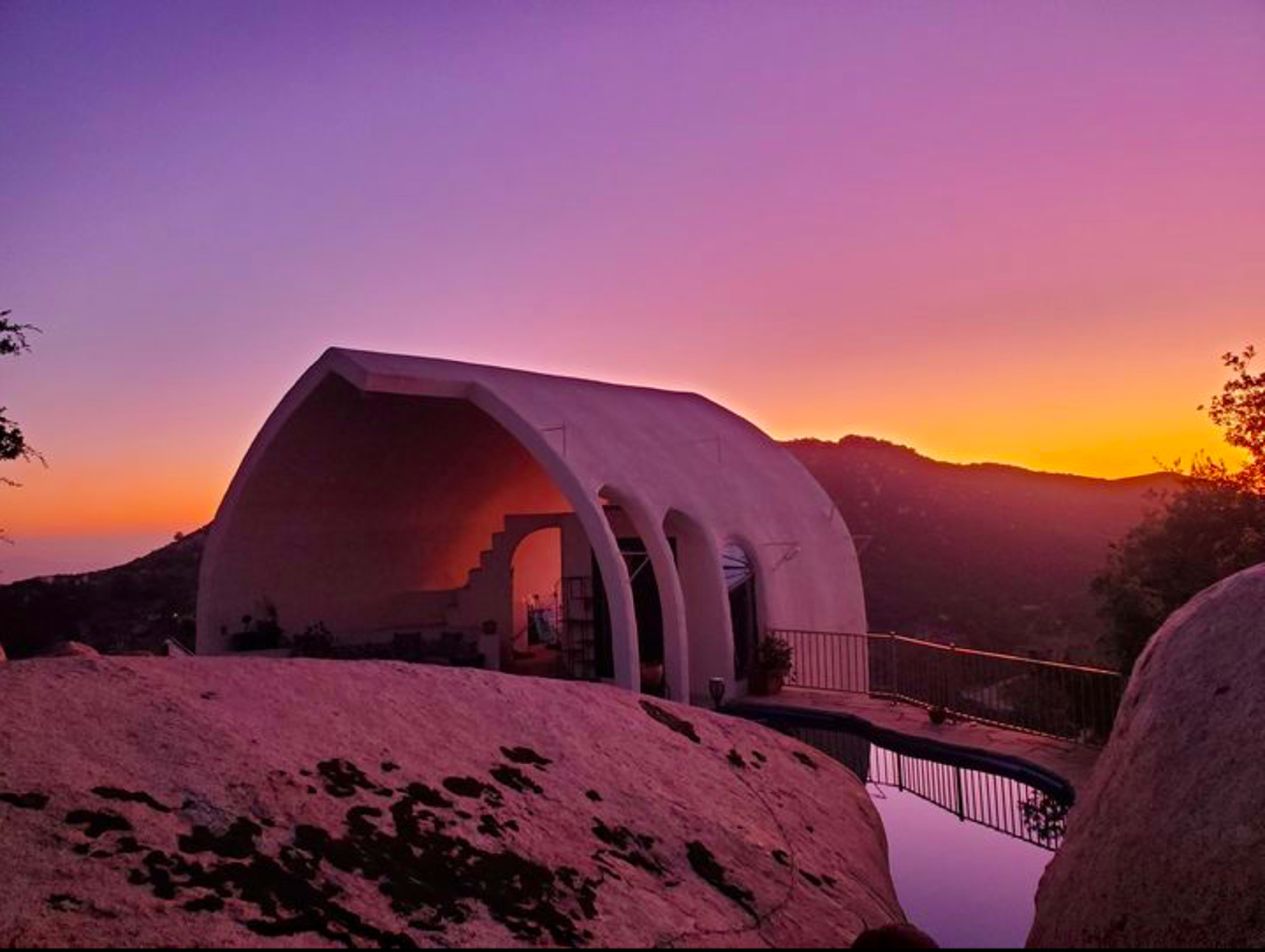 A unique architectural structure sits on rocky terrain, overlooking a colorful sunset.