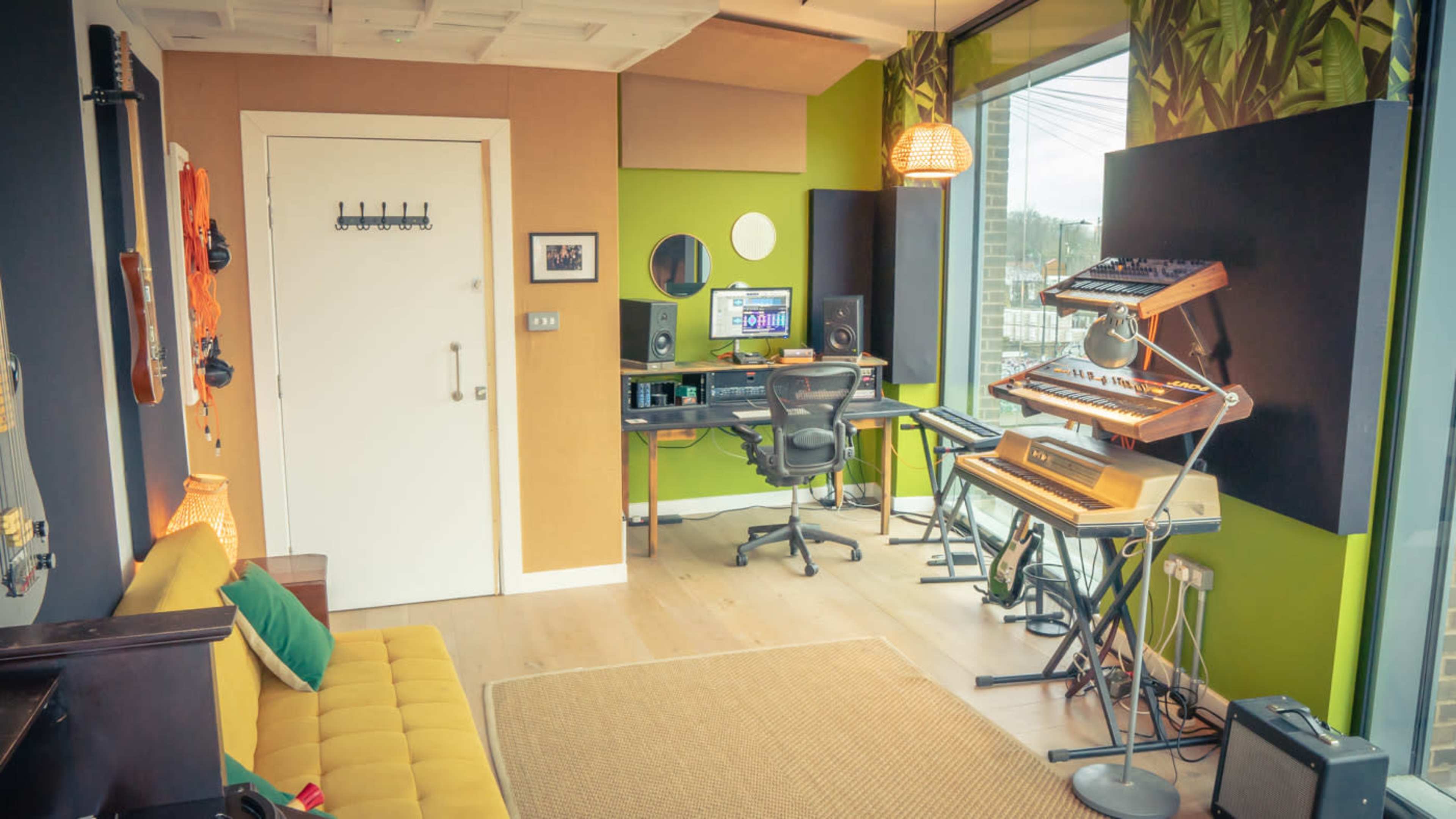 The image shows a small music studio with a desk, computer, and various musical instruments, including a keyboard and synthesizer, set against a modern decor featuring large windows and green walls.