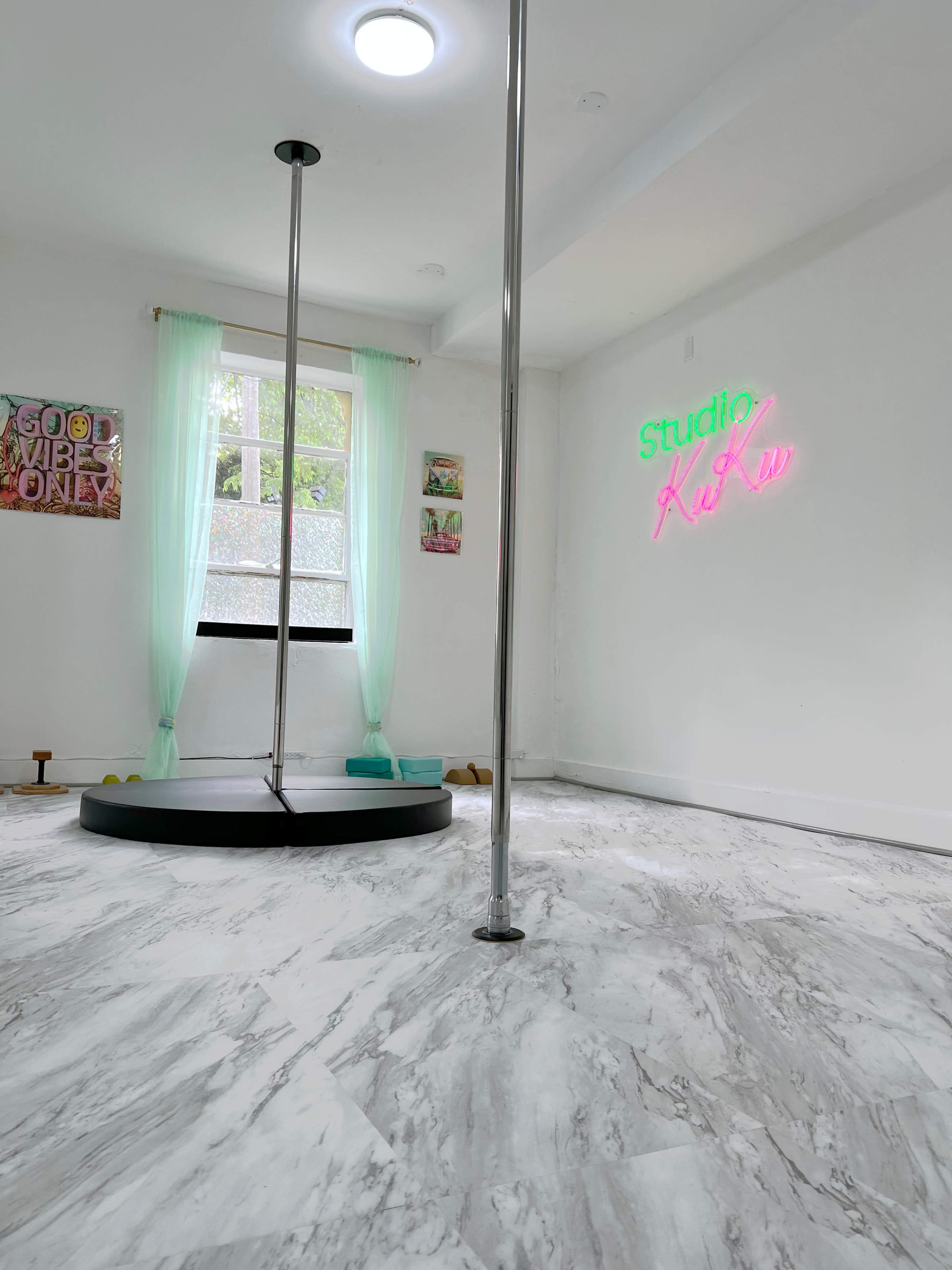 A bright, minimalist dance studio with a central pole, neon signage on the wall, and large windows allowing natural light.