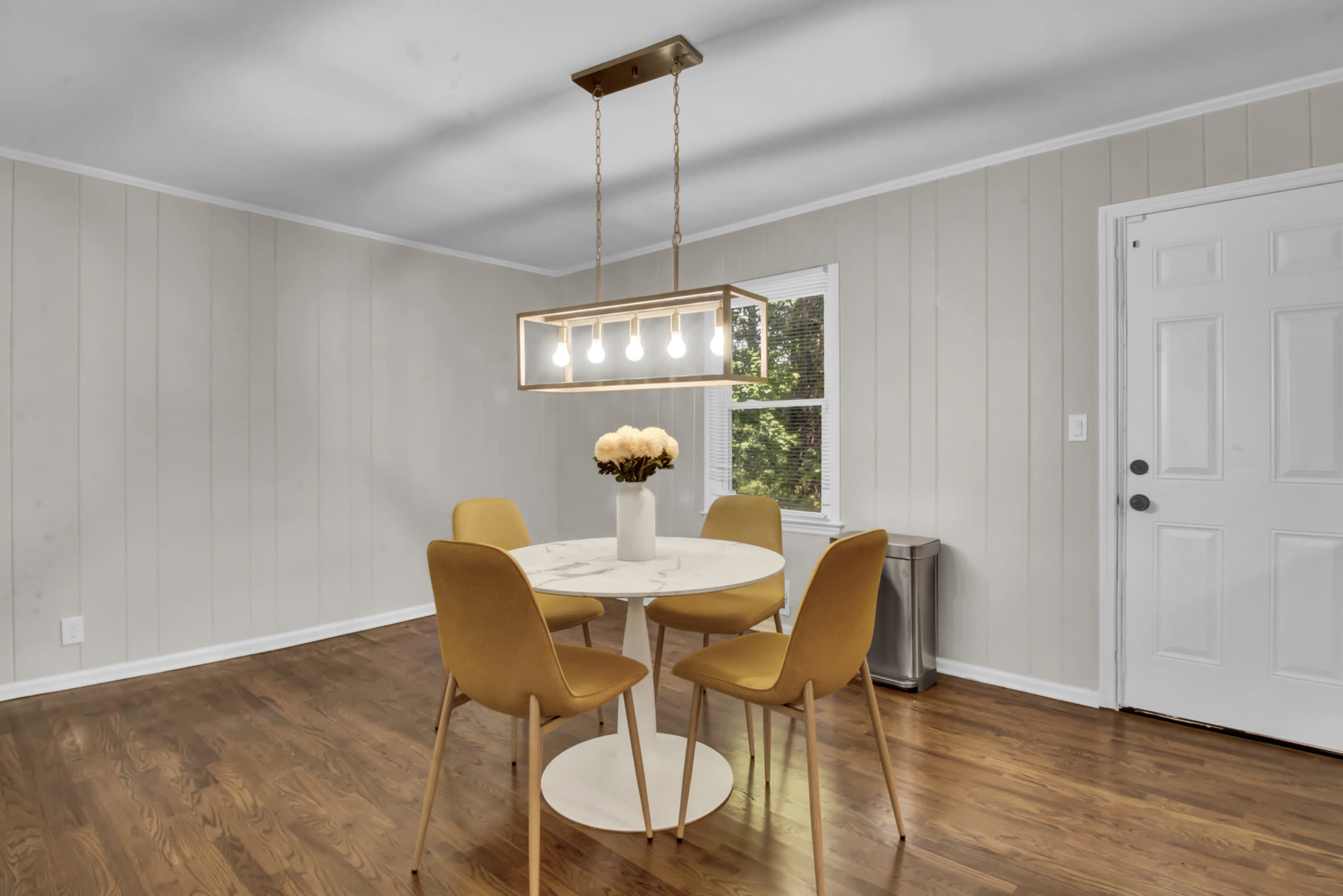 A round table with four yellow chairs is situated in a well-lit dining area with a window and a door.
