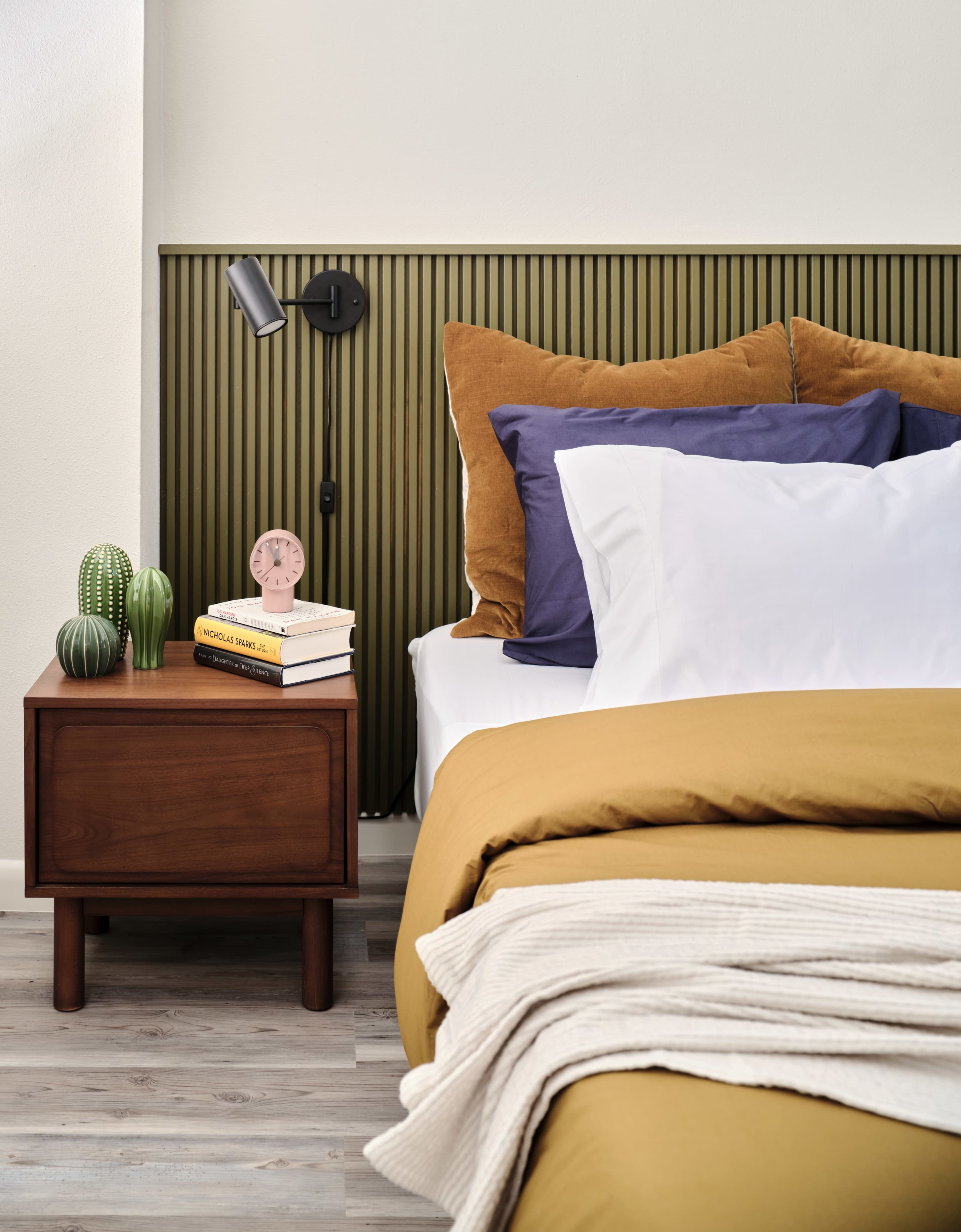 The image shows a modern bedroom with a neatly made bed featuring an assortment of colored pillows, a wooden nightstand with books and a clock, and a wall-mounted lamp above the bed.
