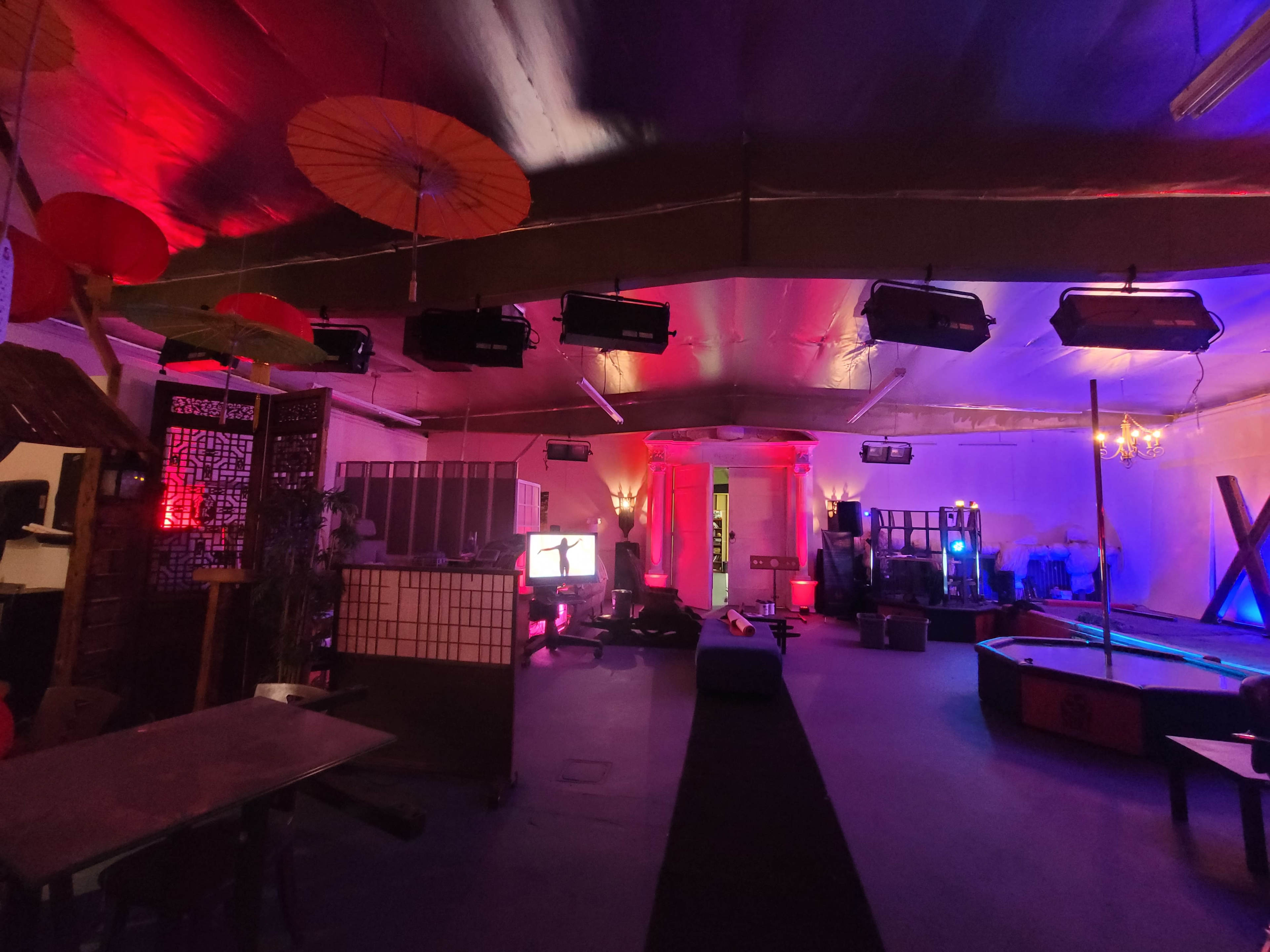 An indoor venue with colorful lighting, featuring decorative elements like umbrellas and screens, and various seating arrangements.