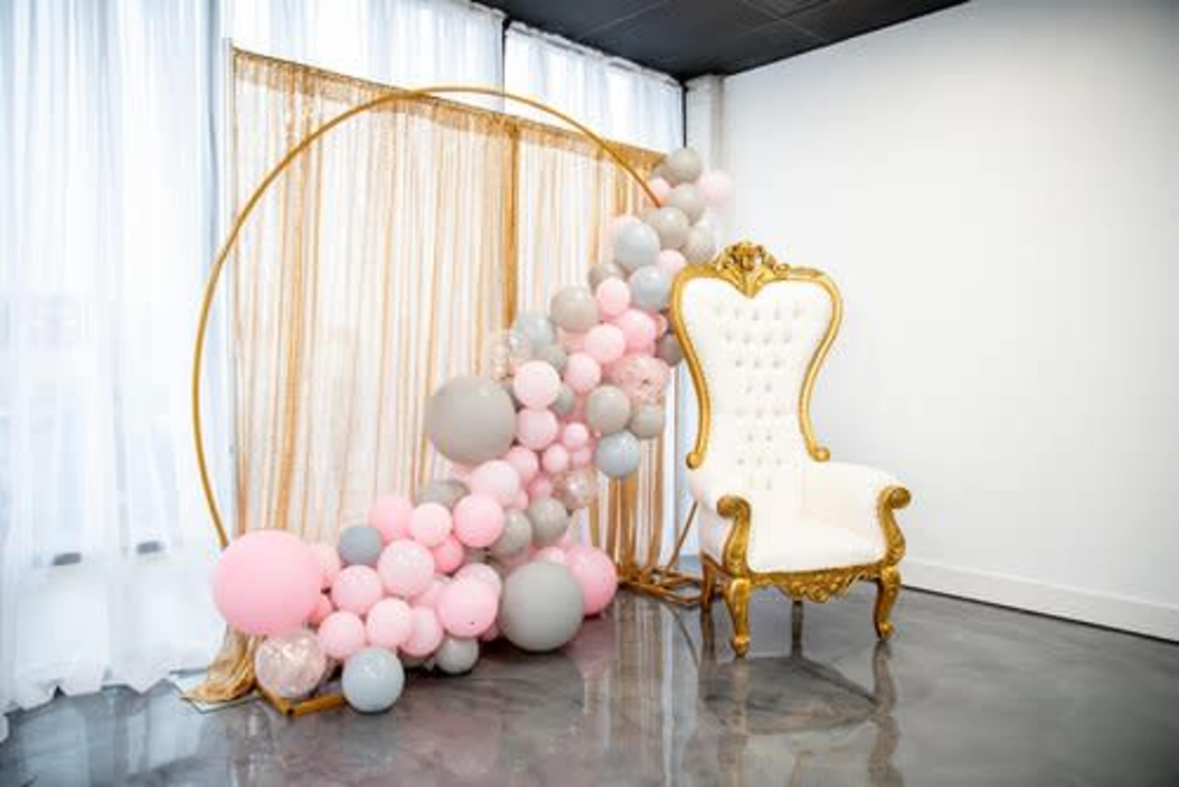 A decorative setup with a golden arch draped in sheer fabric, adorned with a colorful arrangement of balloons, next to an elegant white chair with gold detailing.