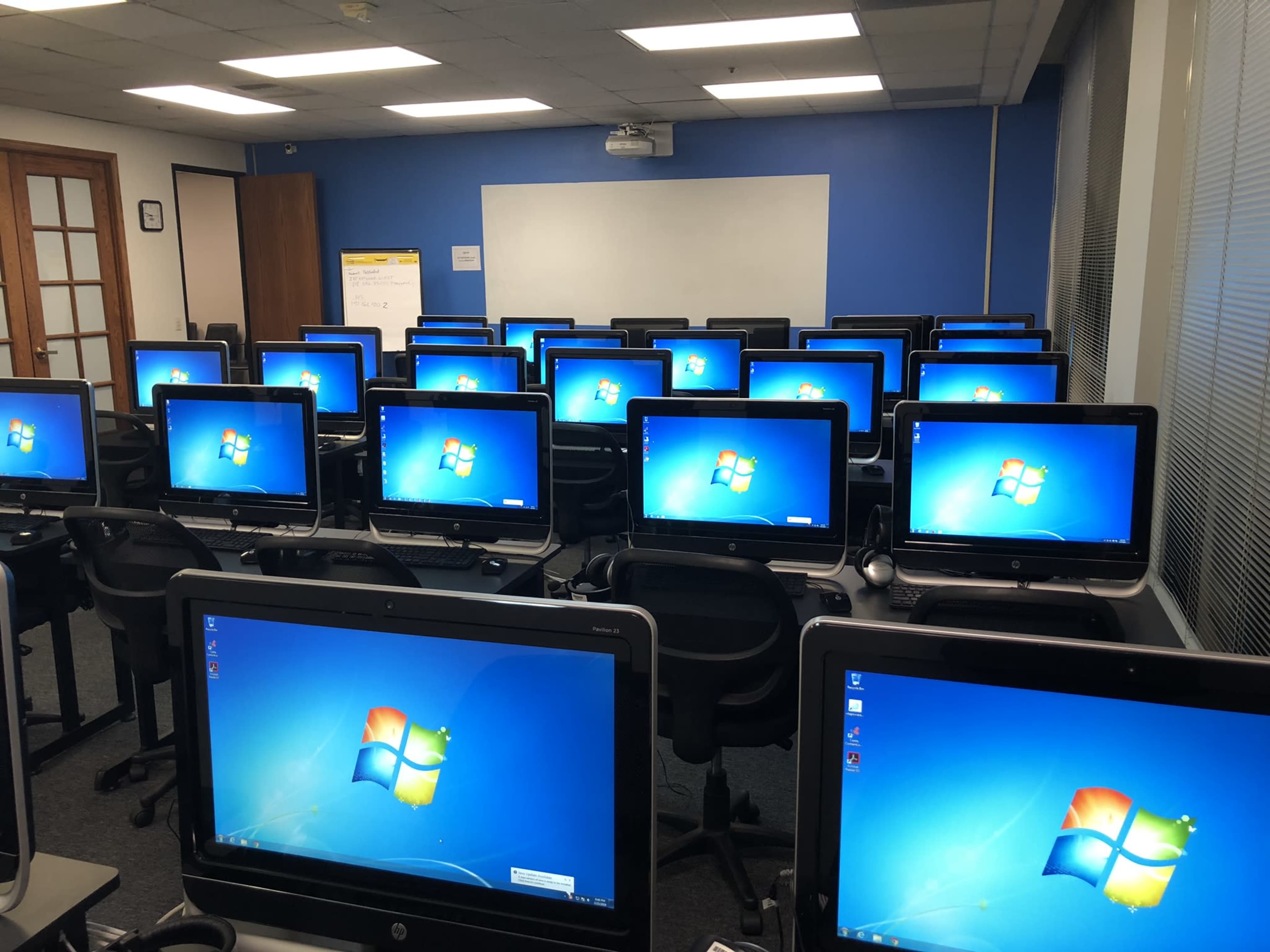 The image shows a classroom with rows of desktop computers, each displaying the Windows login screen.