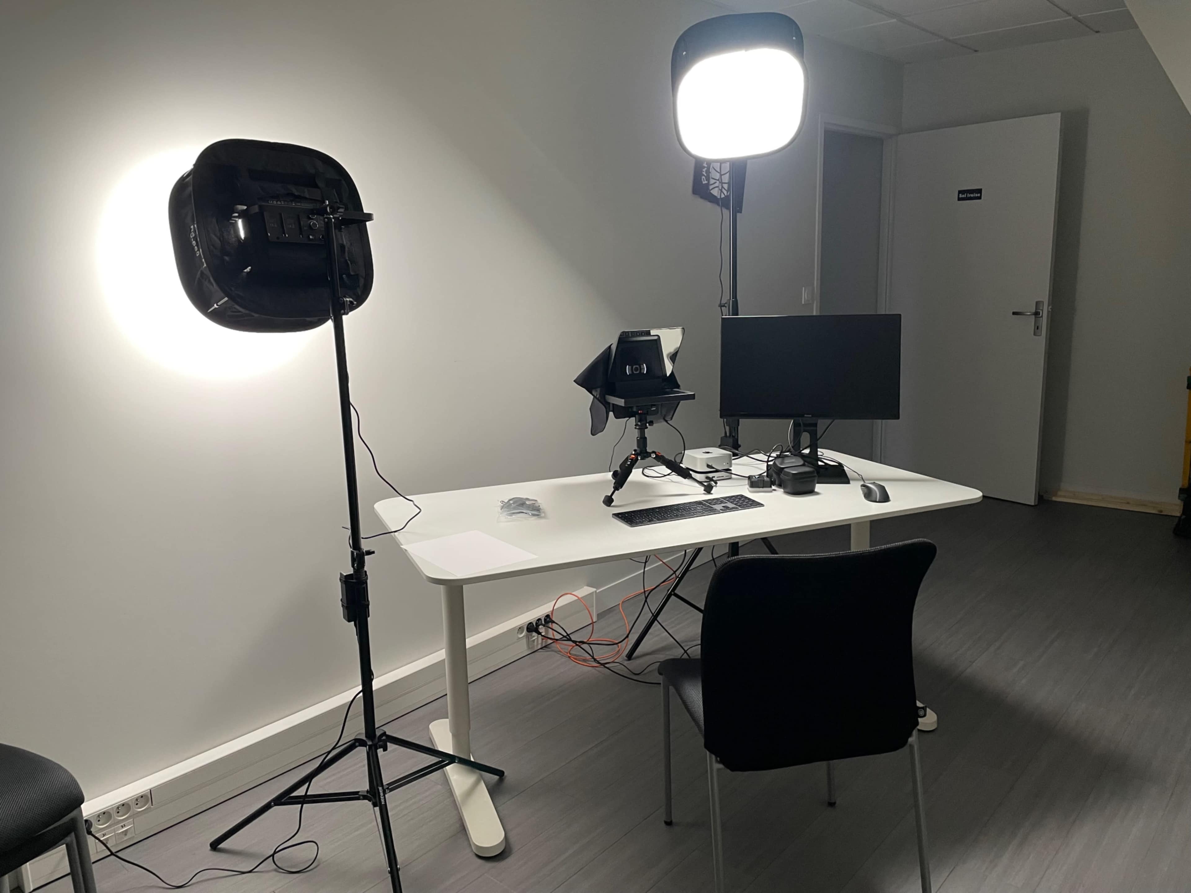 A well-lit workspace features a desk with a computer monitor, camera, and desktop equipment, alongside two lighting setups.