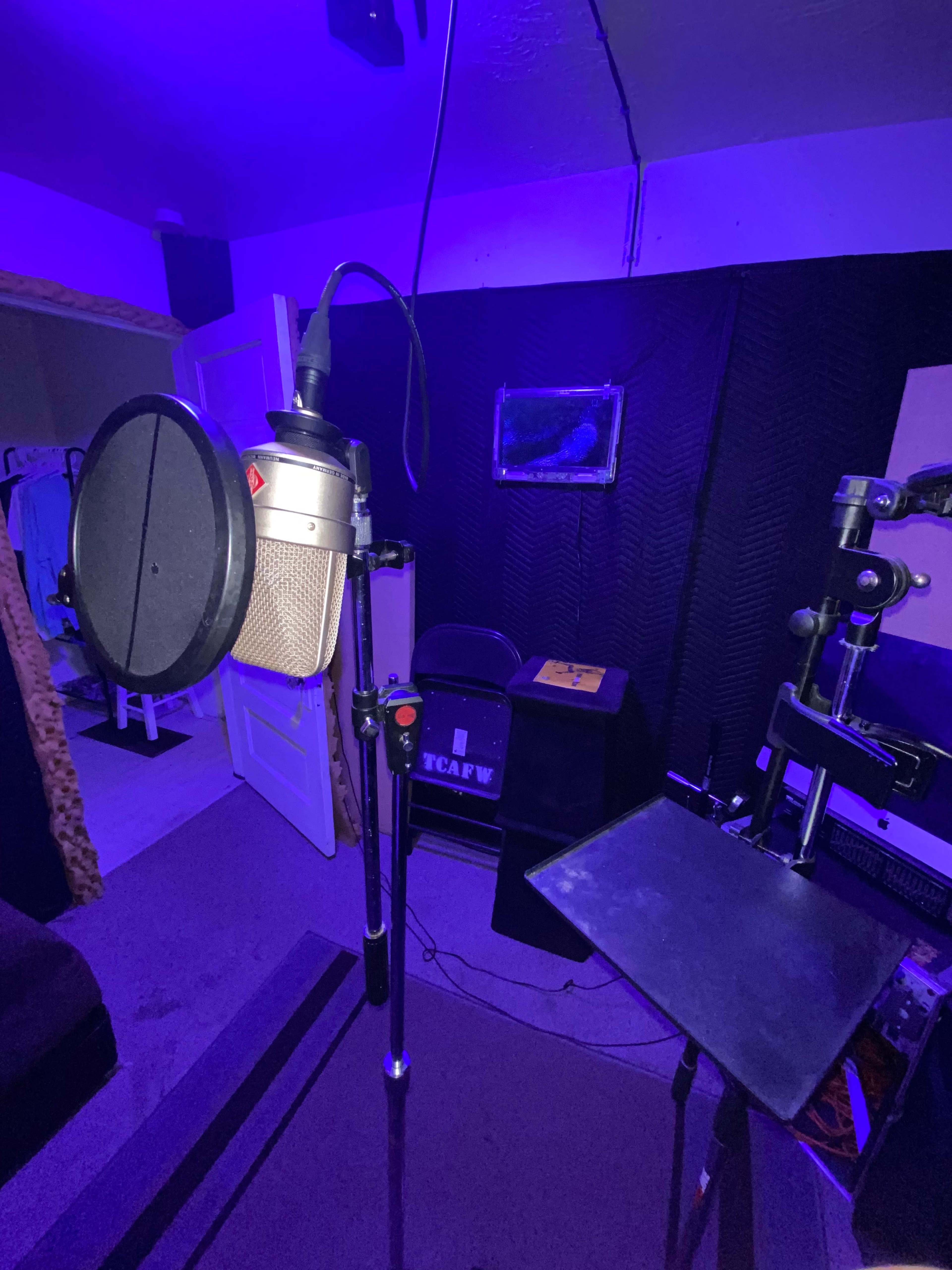 A microphone is positioned on a stand in a dimly lit recording studio with purple lighting and soundproofing panels.