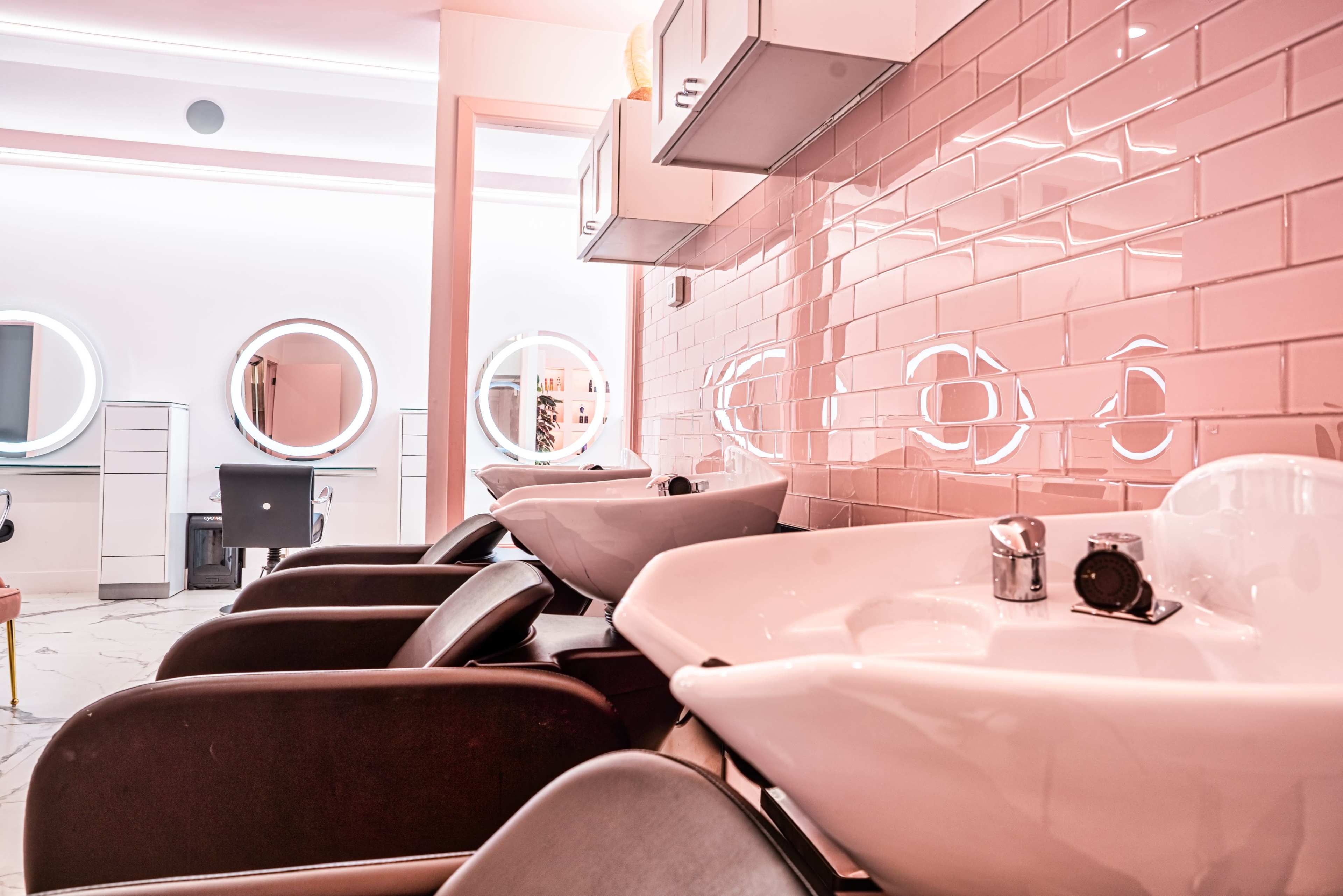 A modern hair salon interior with several salon chairs and wash basins lined against a pink-tiled wall.