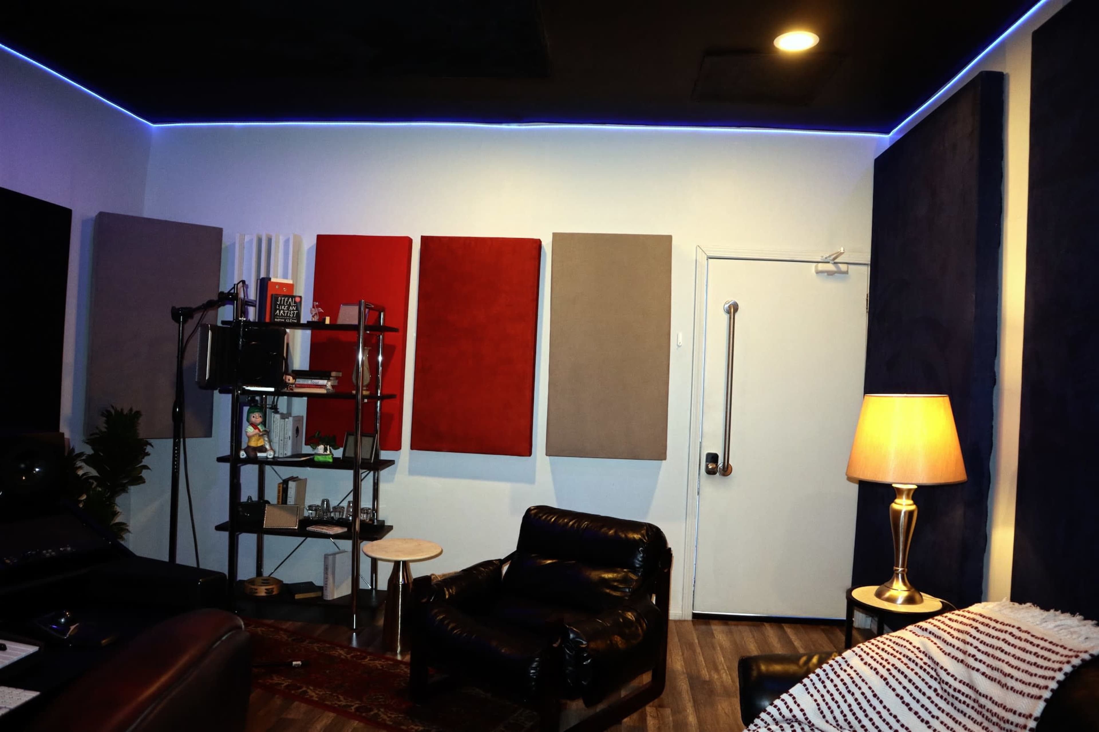 The image shows a music studio featuring soundproofing panels on the walls, a black shelving unit with equipment, a small table, and a leather armchair, all illuminated by LED lighting.