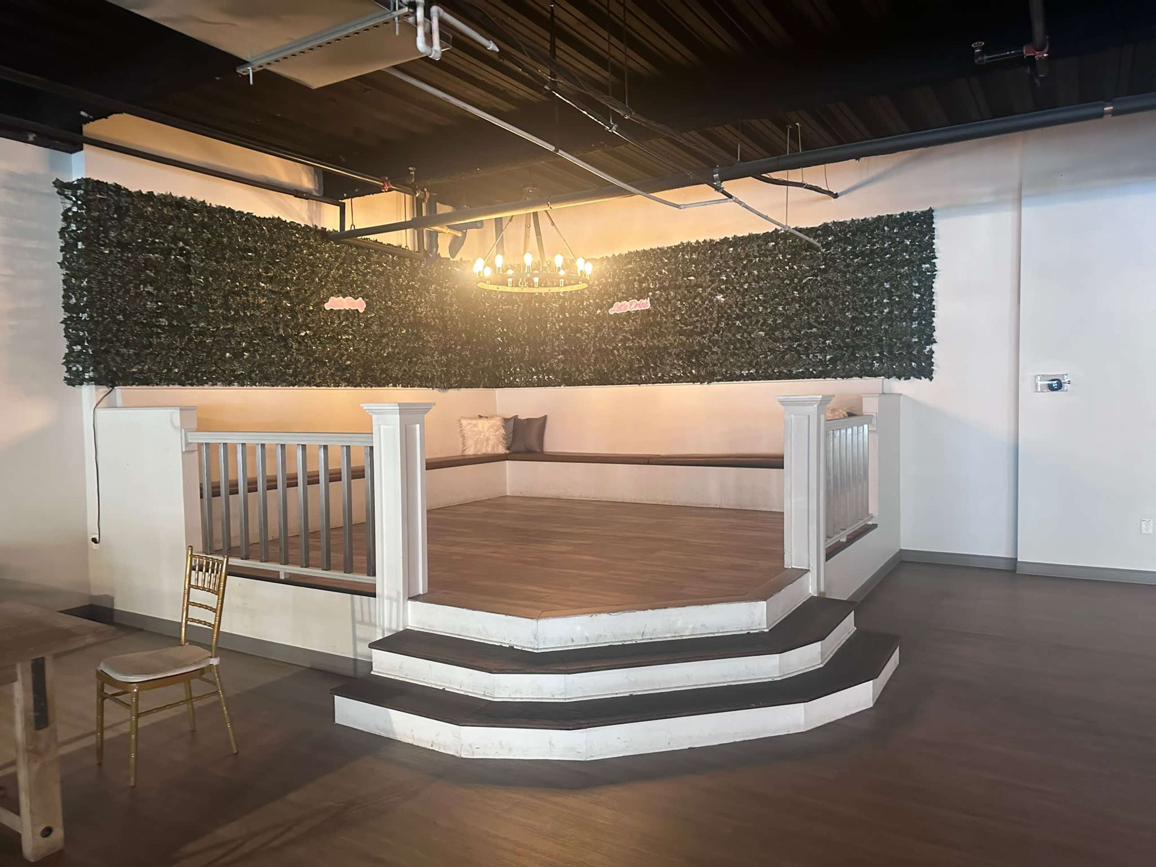 The image features a stage area with a raised platform, surrounded by a green foliage wall and illuminated by a chandelier.