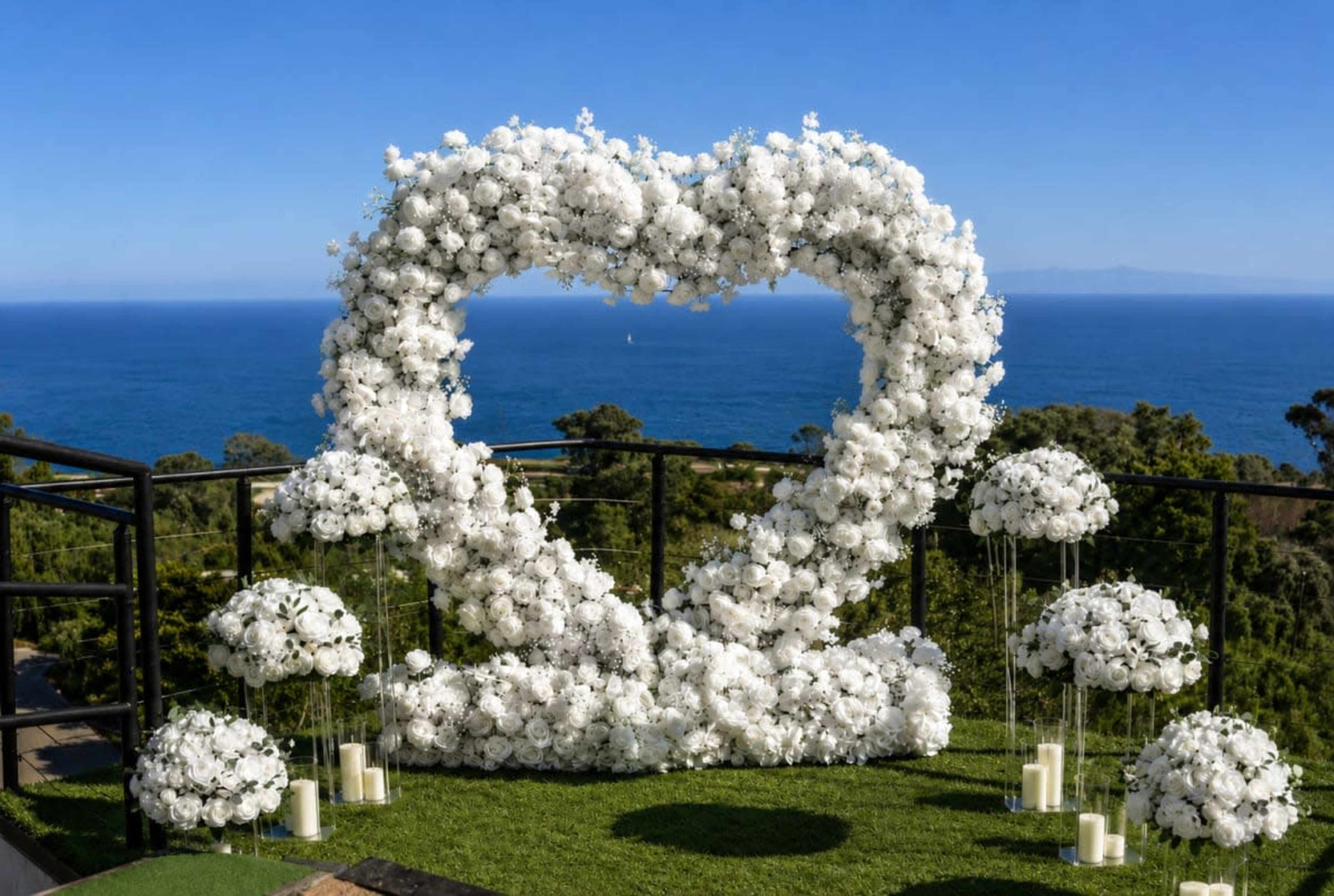 A heart-shaped floral installation made of white flowers is set up on a balcony overlooking the ocean, accompanied by smaller floral arrangements and candles.