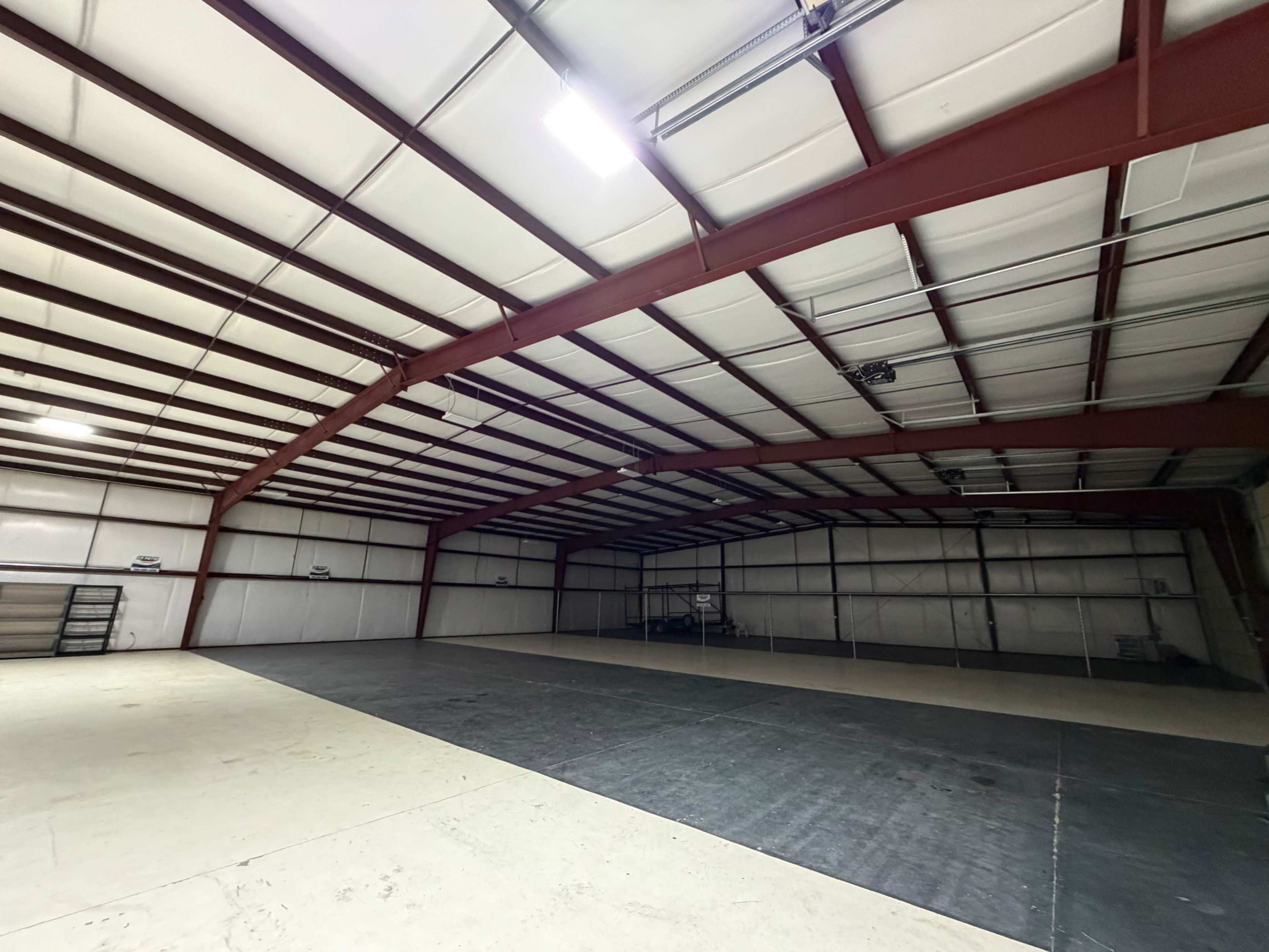 The image shows the interior of a large industrial building with a high ceiling and metal beams, featuring an open, unoccupied space on the floor.