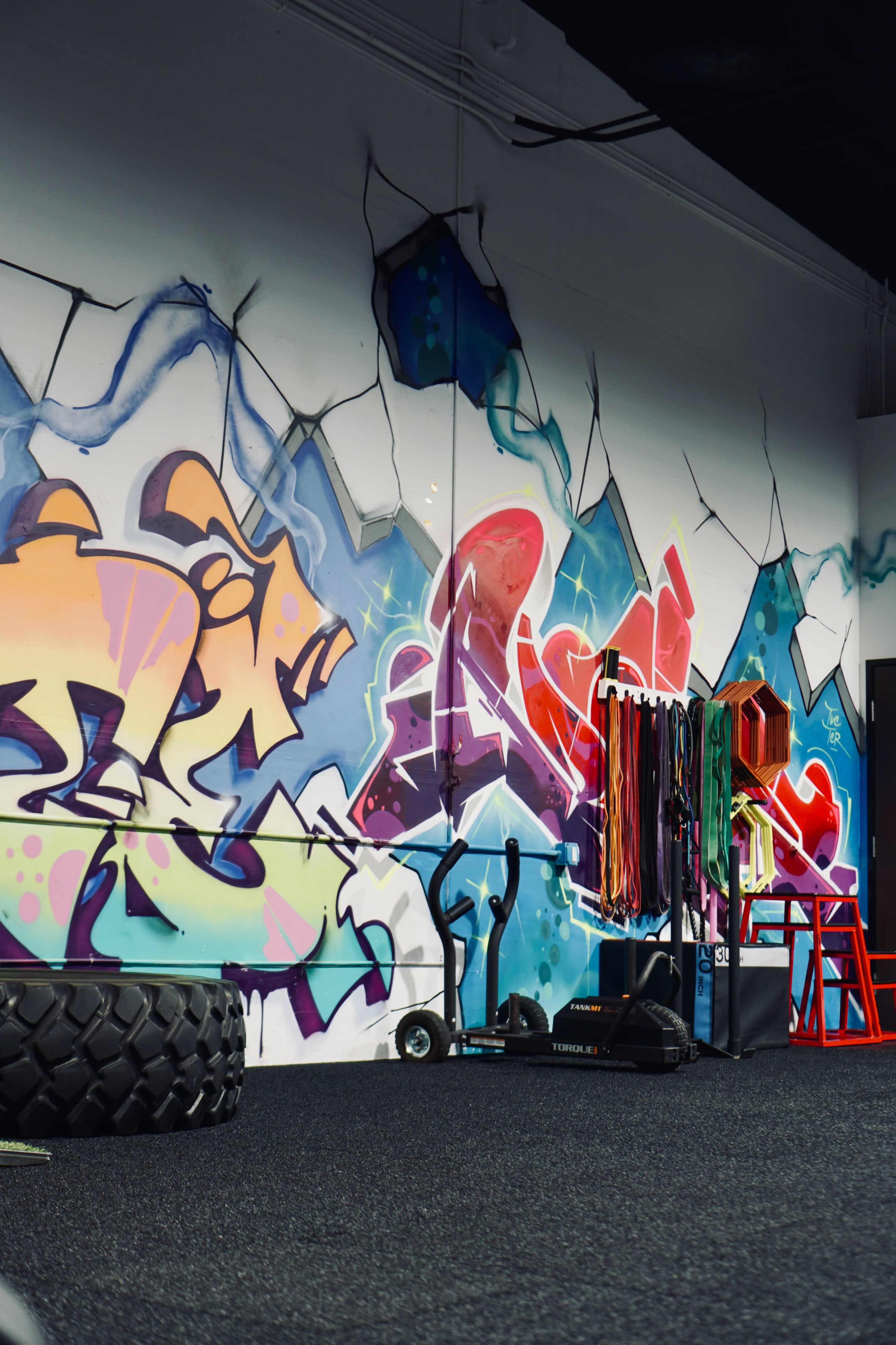 The image shows a gym interior with colorful graffiti on the wall and exercise equipment, including resistance bands, against a black rubber floor.