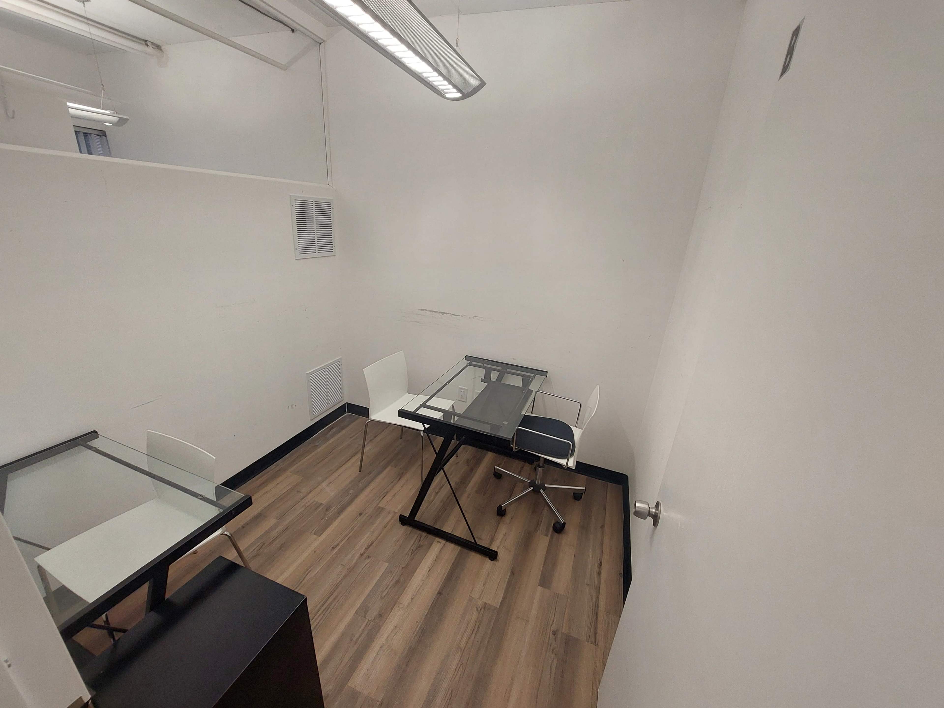 The image shows a small, empty office space with two desks and two chairs on a wooden floor.