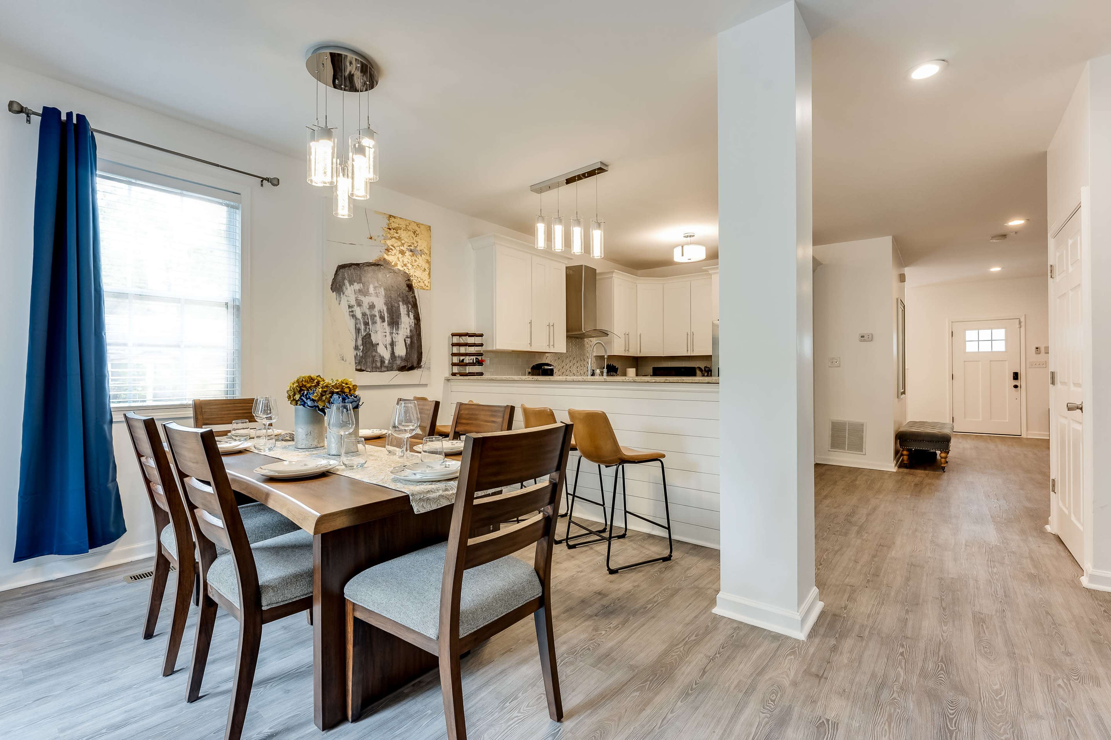 A dining area with a table set for six, surrounded by wooden chairs, leads to an open kitchen featuring modern appliances and a bar with stools.