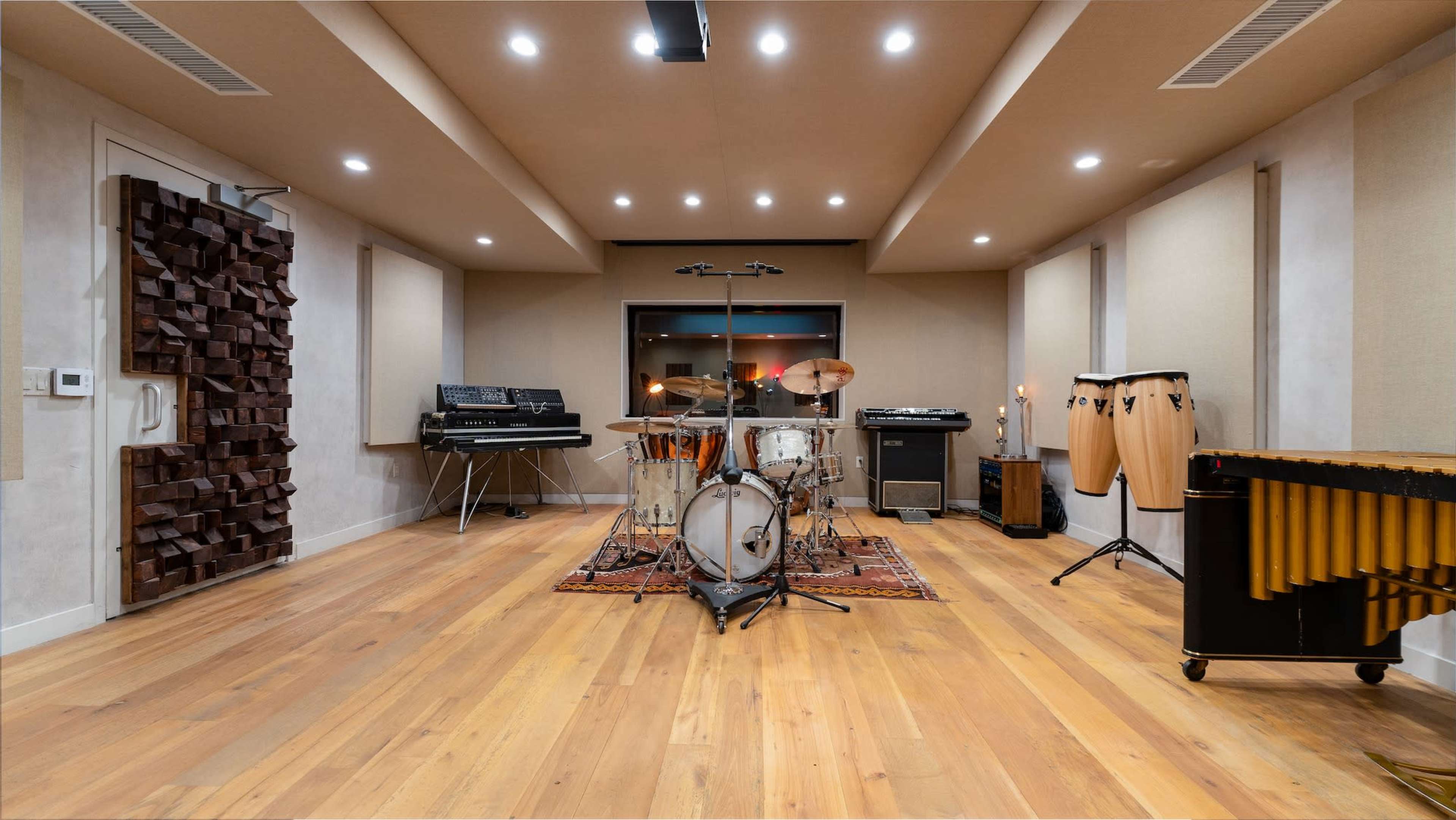A well-equipped music studio features a drum set, percussion instruments, synthesizers, and soundproof walls with wooden accents.