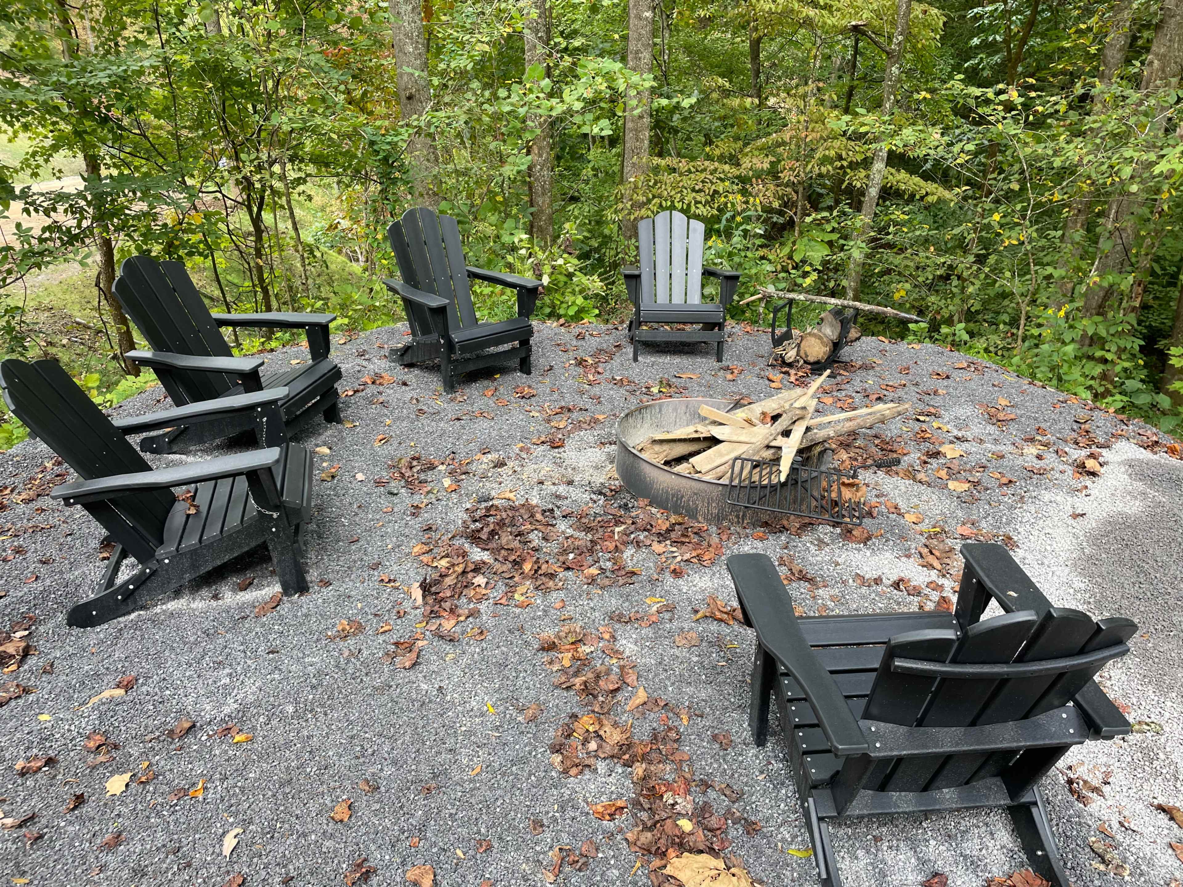 A circle of six black outdoor chairs surrounds a fire pit with firewood in a wooded area.