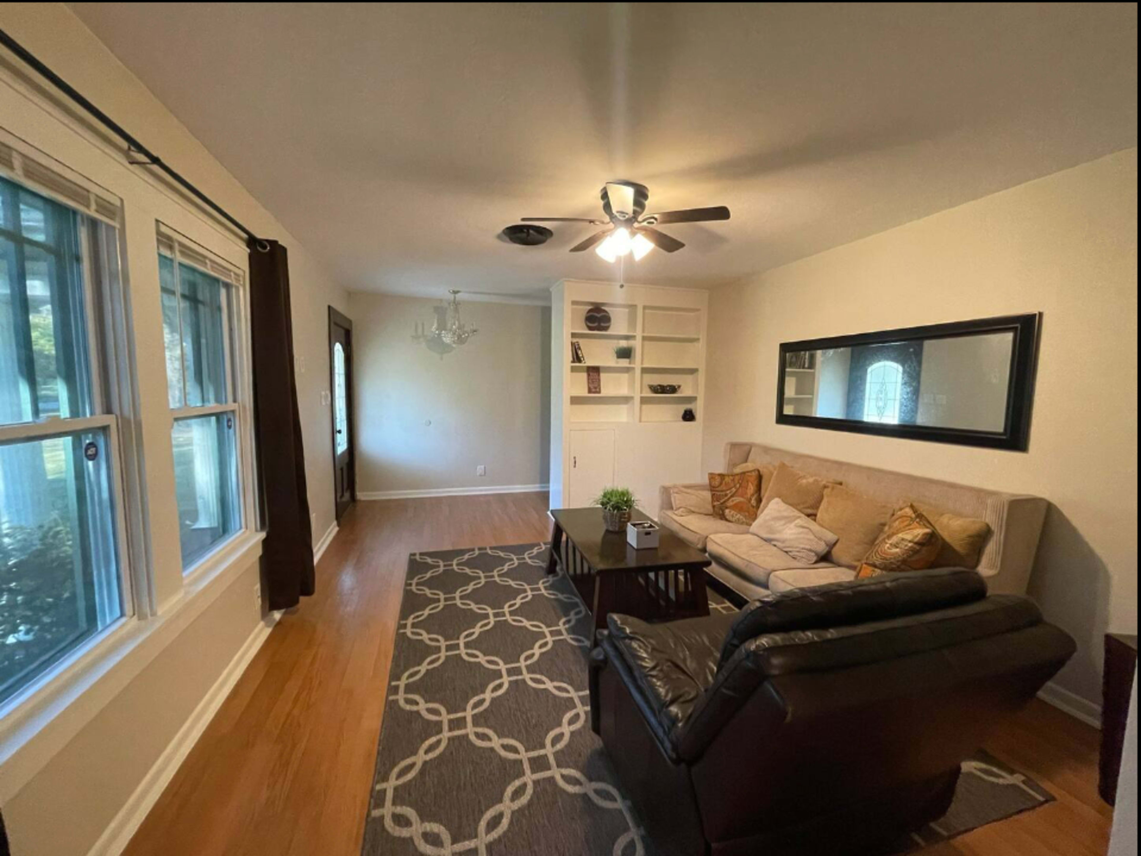 A living room with a beige sofa, a black recliner, a coffee table, and large windows allowing natural light, along with shelves and a chandelier.