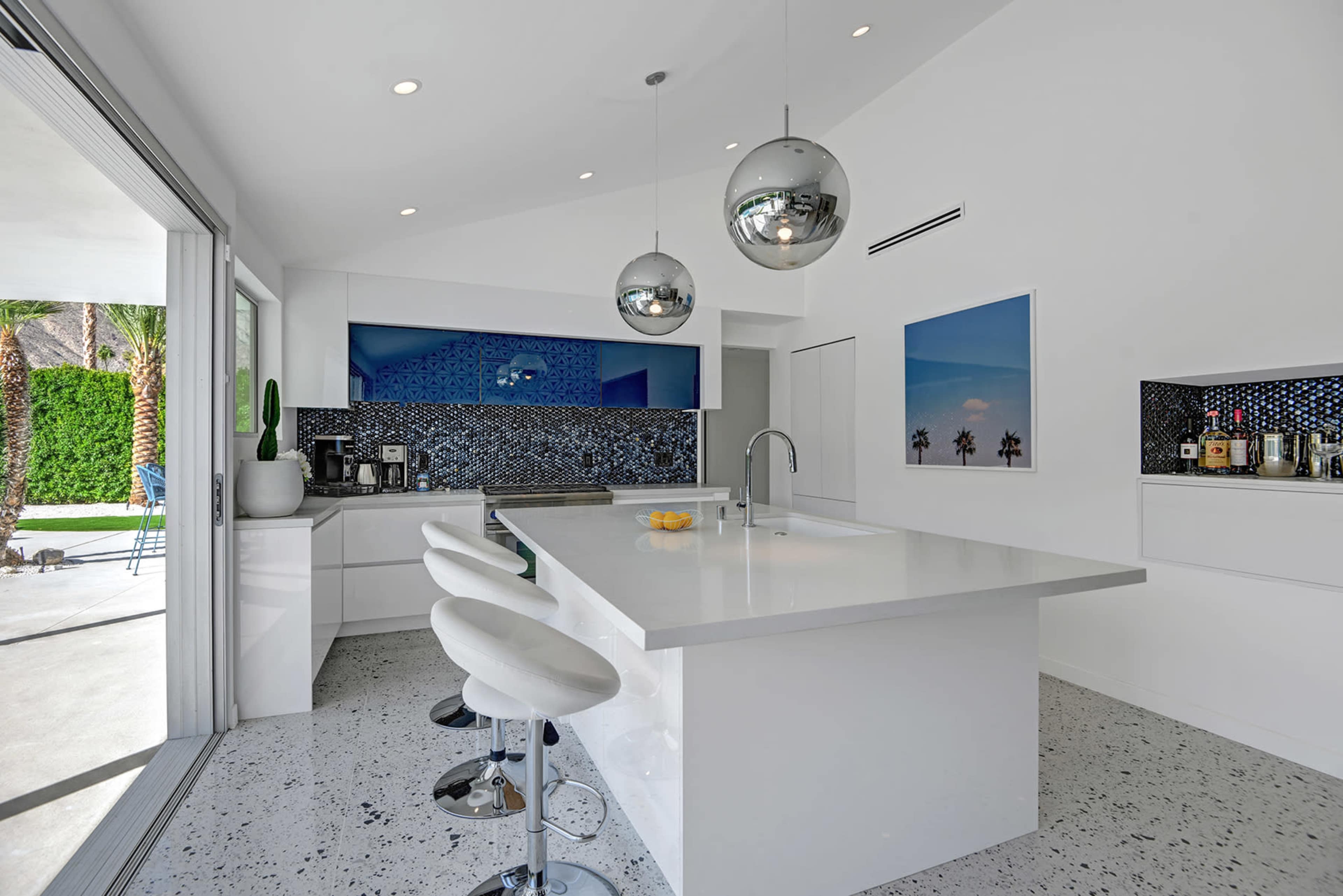A modern kitchen features a large island with white countertops, three white bar stools, and sleek pendant lights, set against blue mosaic tile and large windows overlooking a patio.