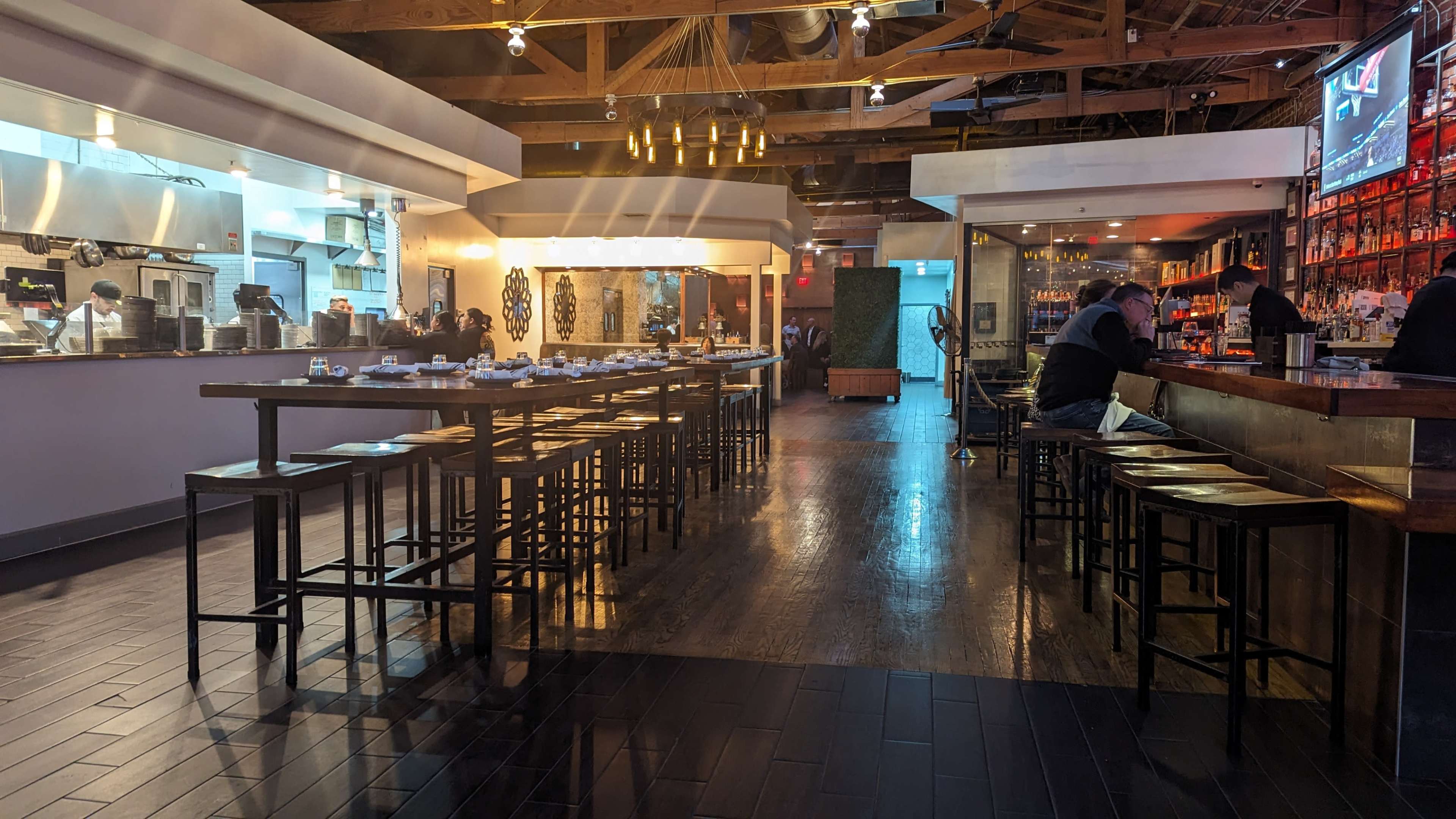 The image shows a restaurant interior with a long bar and dining area, featuring empty stools and tables under warm lighting.