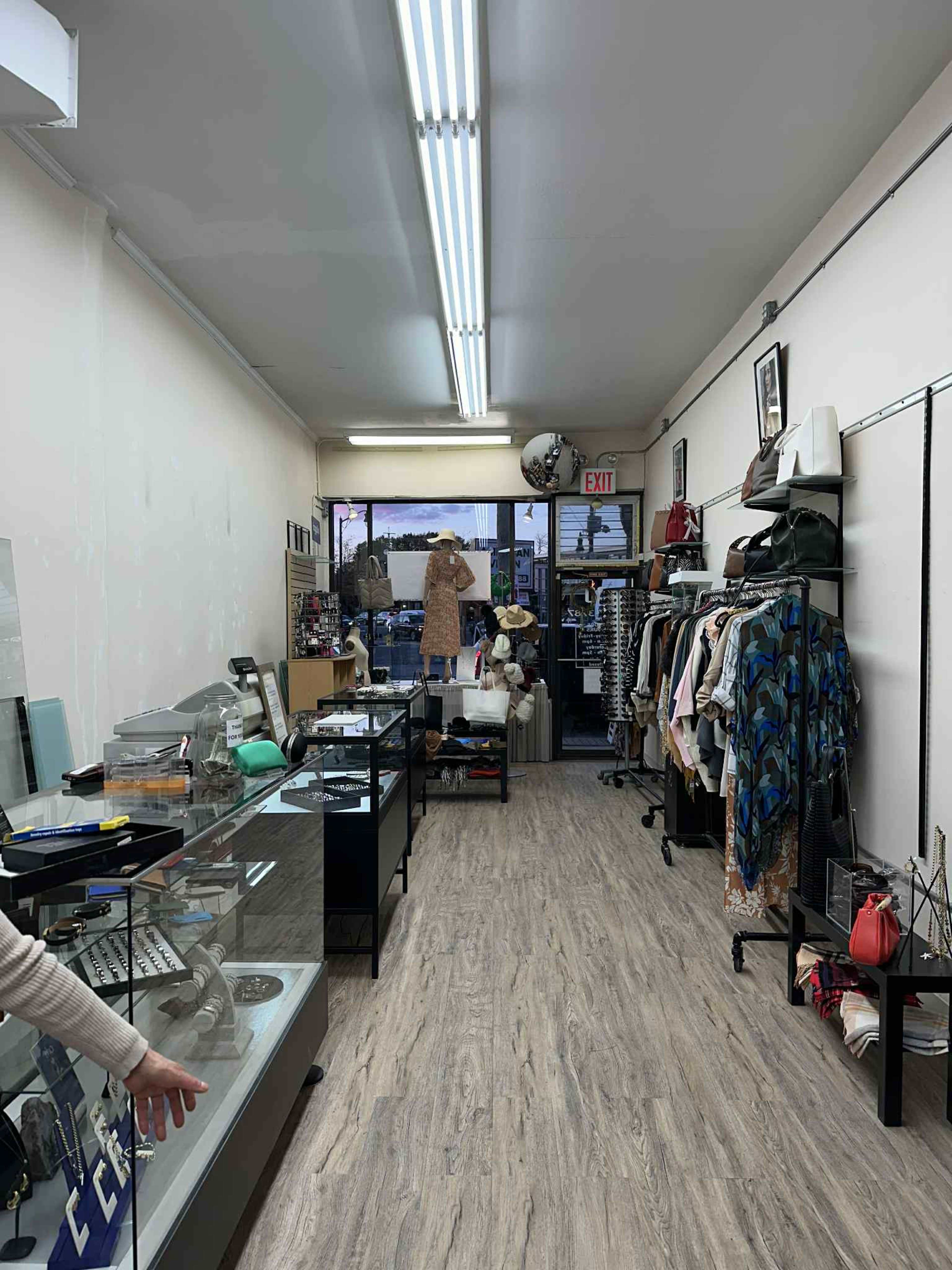 The interior of a thrift shop features racks of clothing, display cases with various items, and a bright, open layout leading to the entrance.