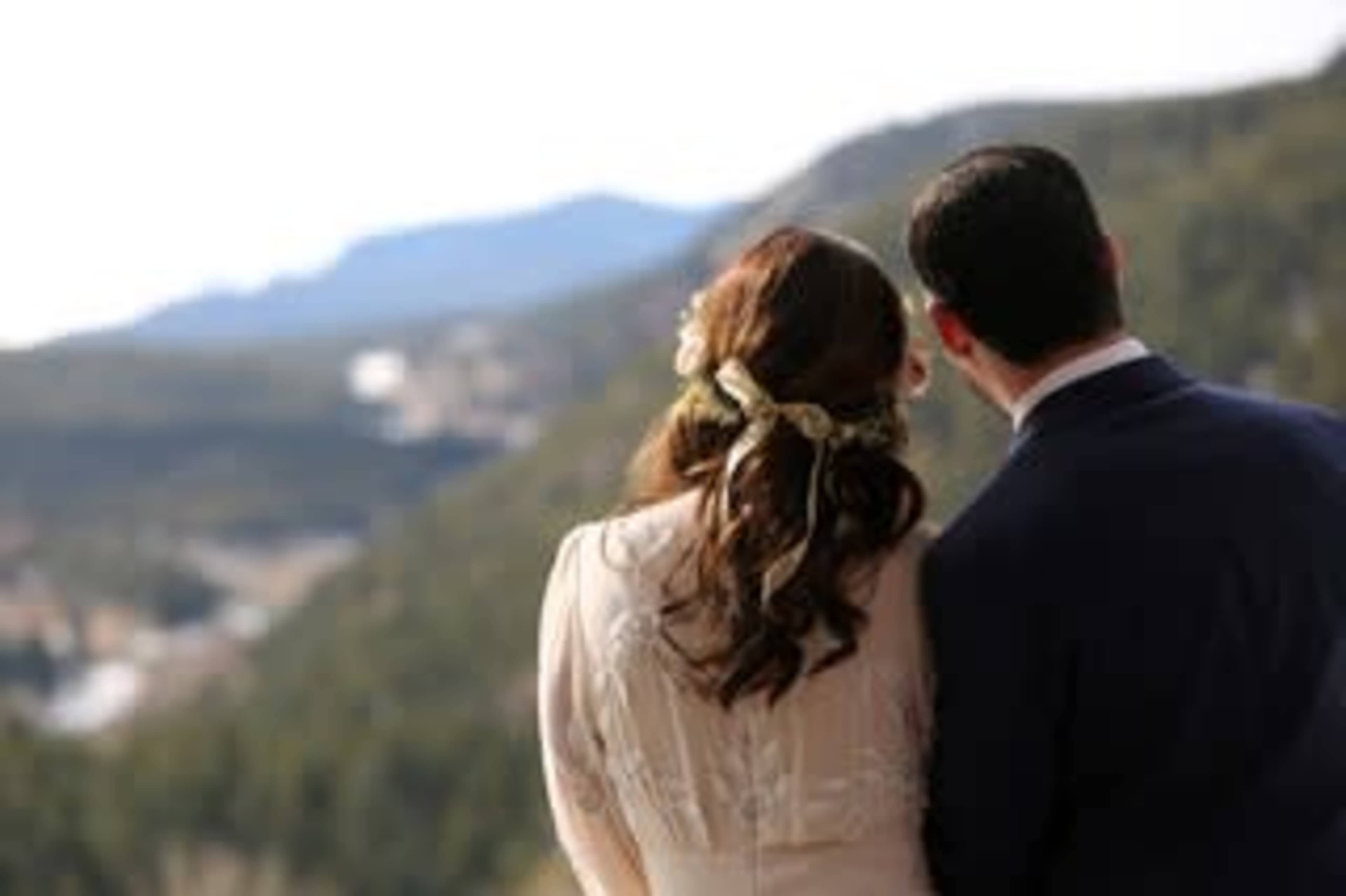 A couple stands together, overlooking a mountain landscape, with the bride's hair adorned with flowers.