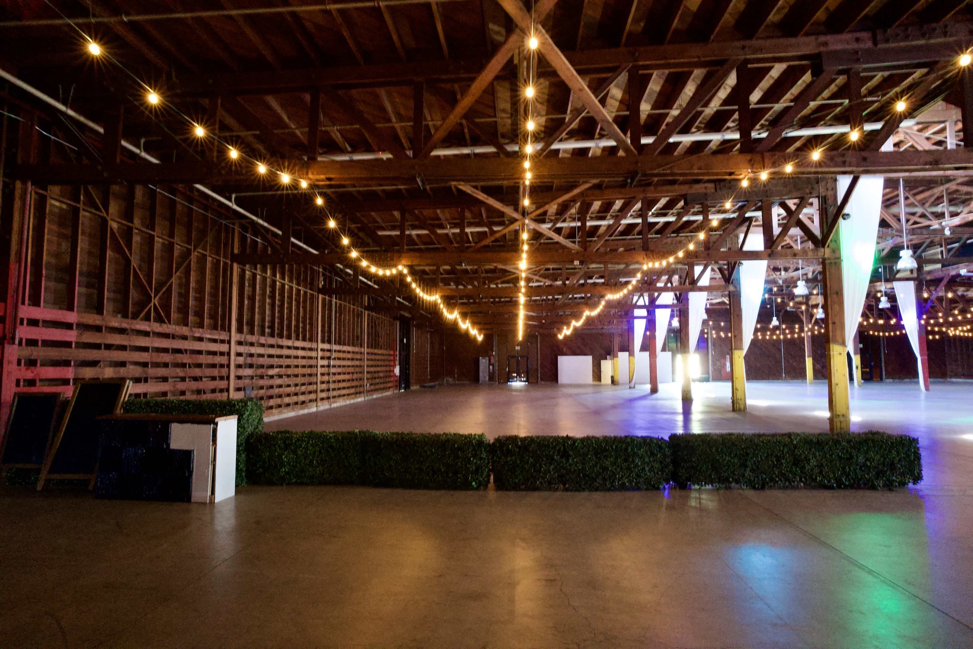 The image shows a spacious event venue with wooden beams, string lights overhead, and a concrete floor, lined with hedges along one side.