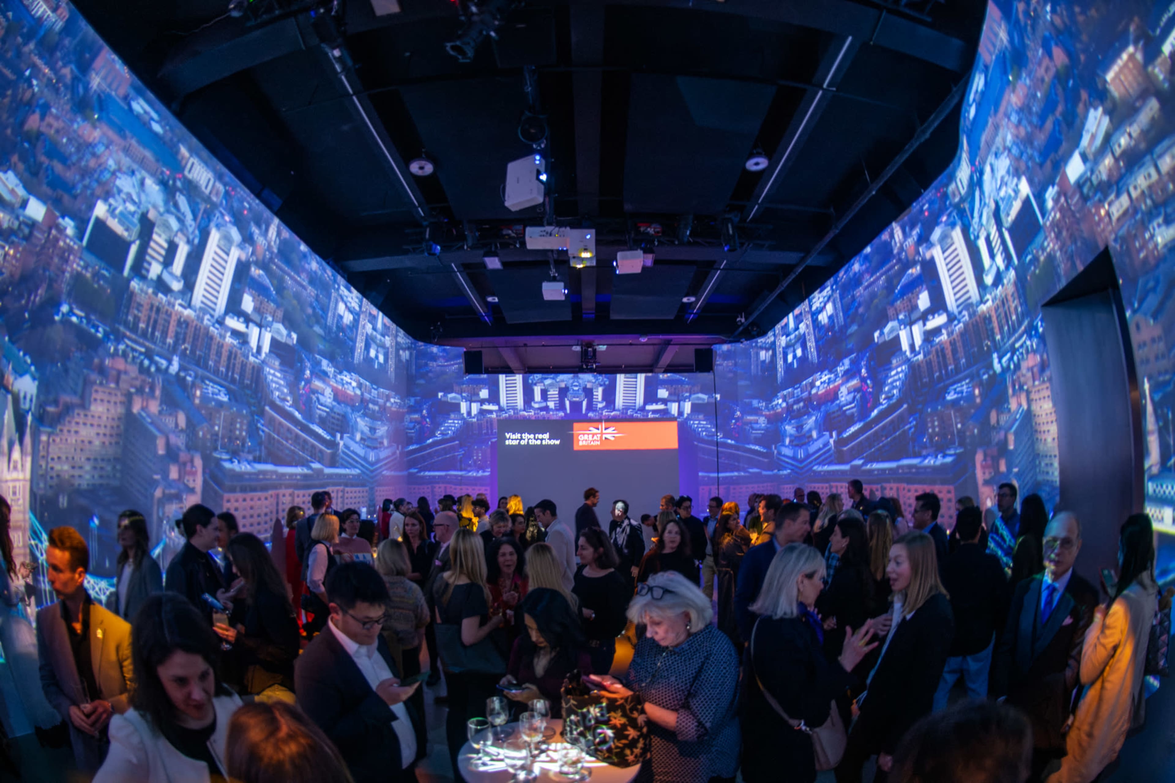 A crowded event space features attendees mingling among projected cityscape visuals on the walls.