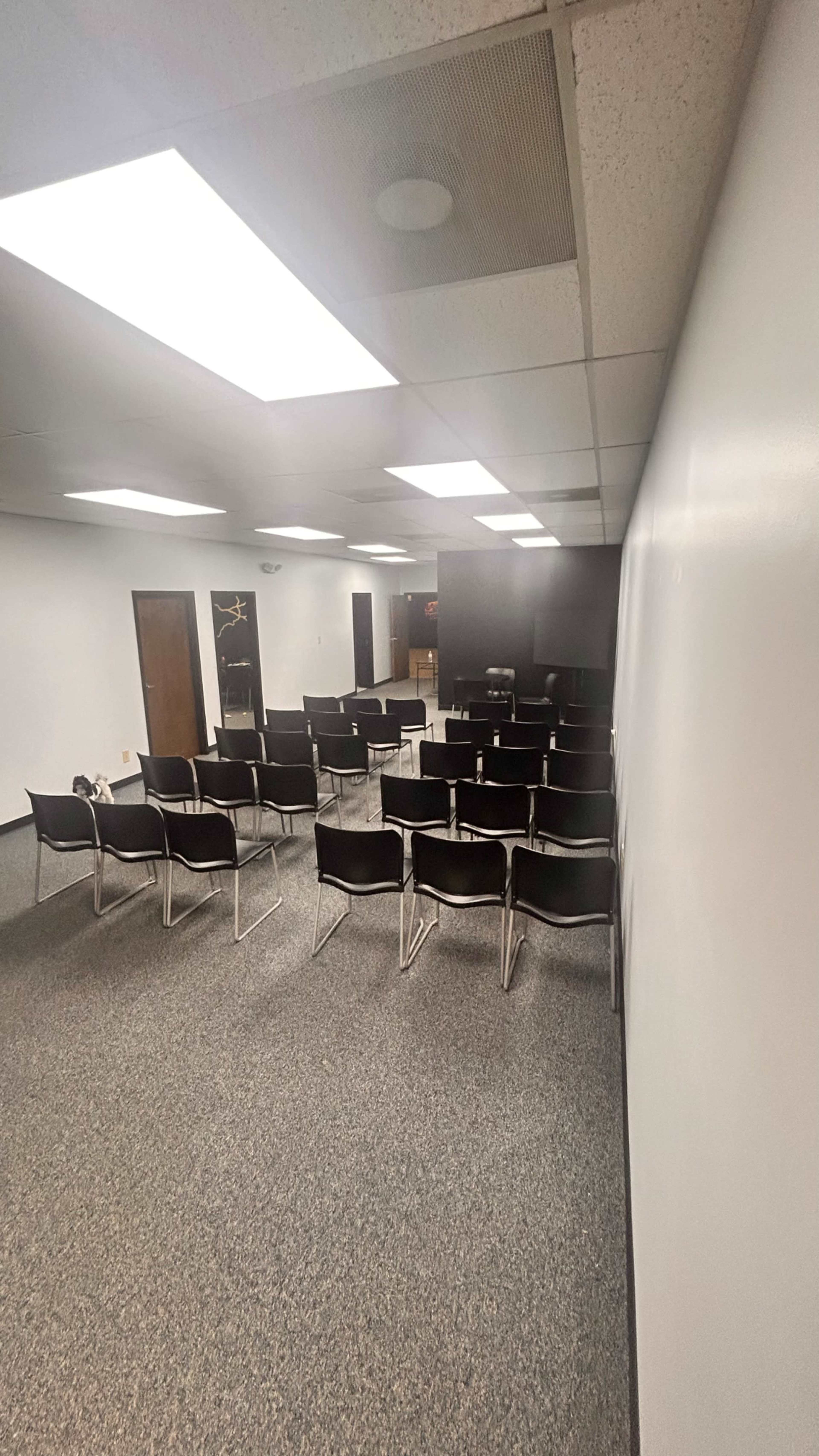 The image shows a conference room with several rows of black chairs arranged facing a blank wall.