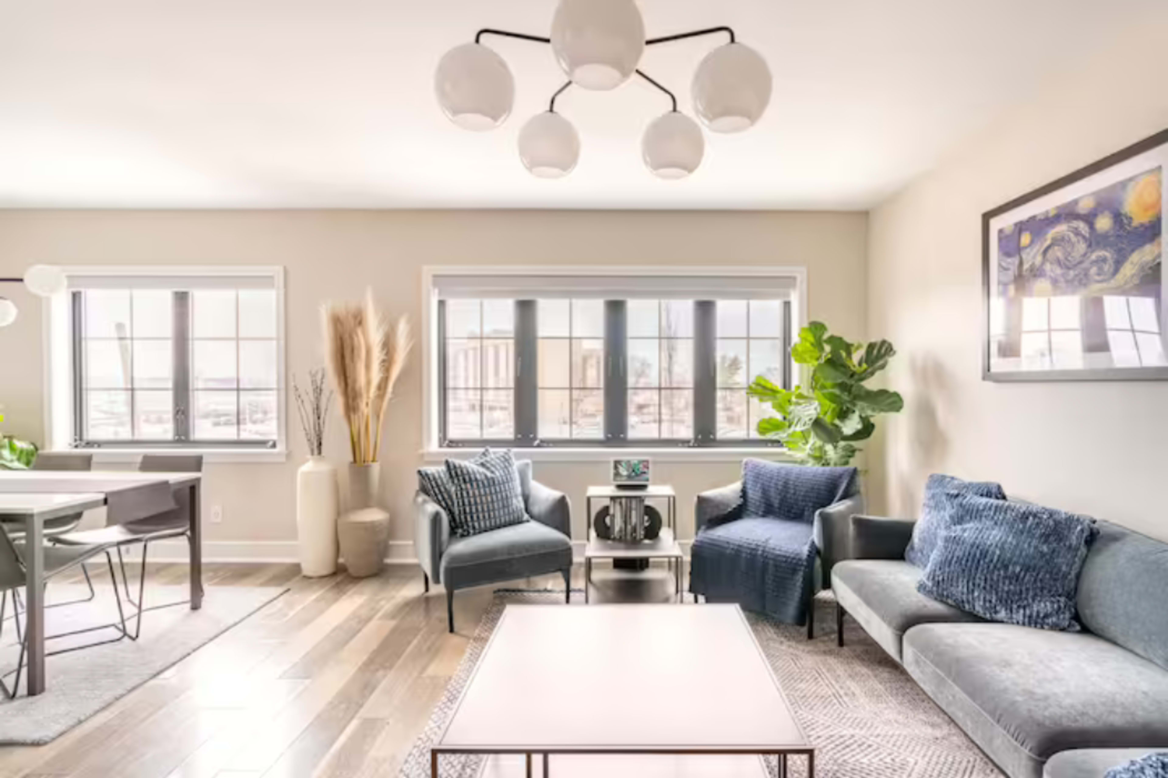 The living room features a modern design with a coffee table, two upholstered chairs, and a sectional sofa, complemented by large windows and decorative plants.
