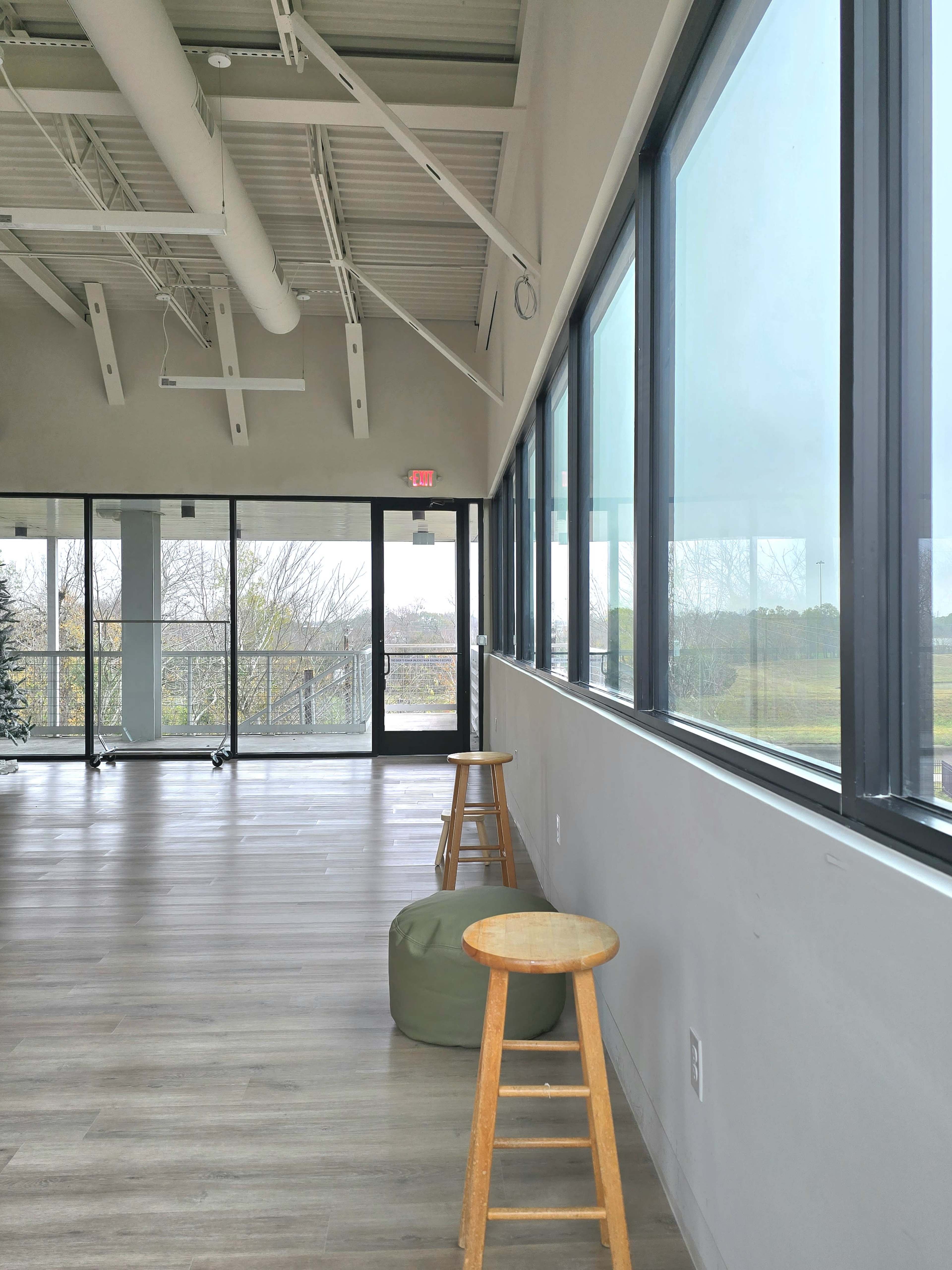 The image shows an empty room with large windows, wooden flooring, and two stools, one wooden and one green, positioned along a wall.