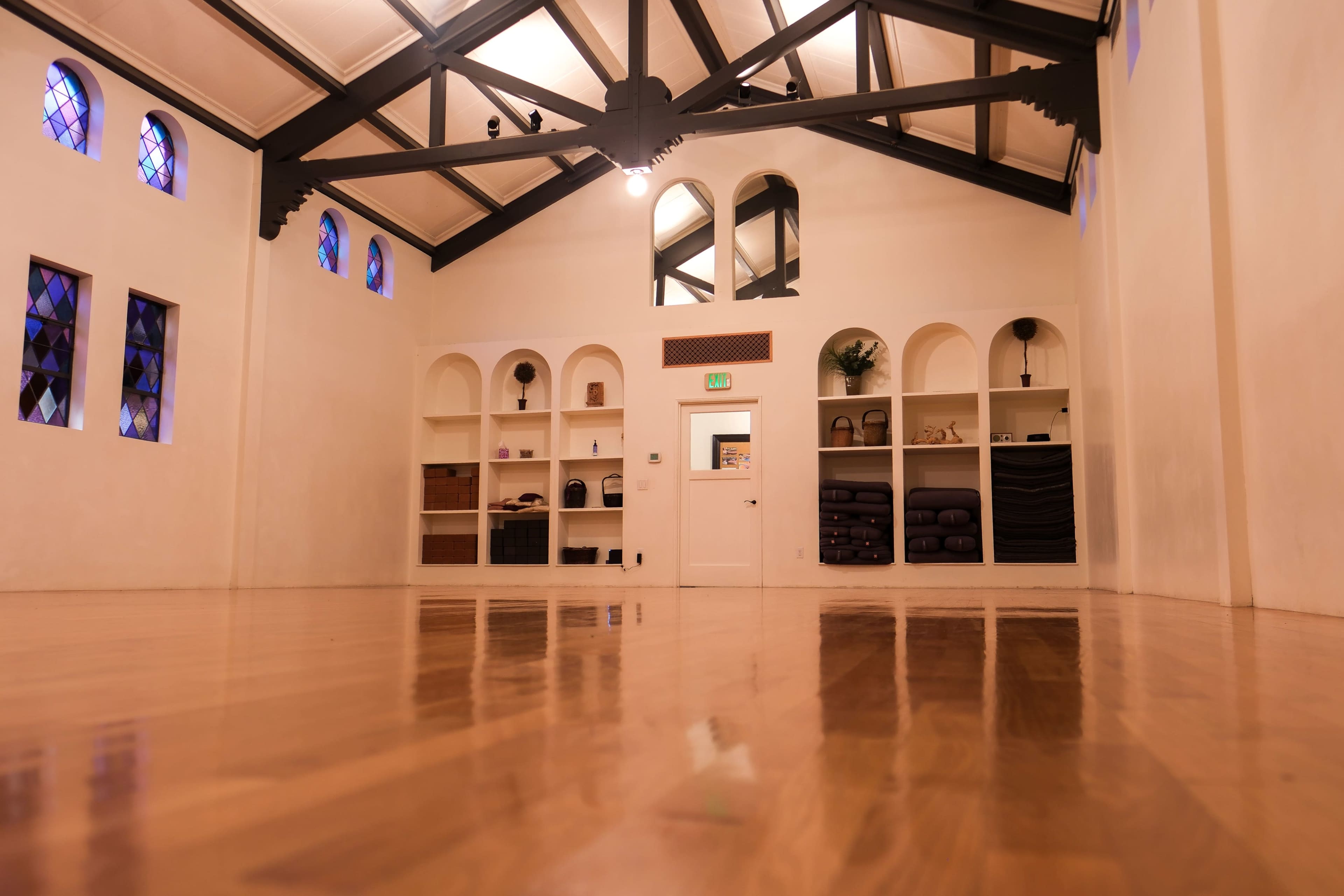 The image depicts a spacious, brightly lit room with wooden flooring, high ceilings, and shelves displaying various items.
