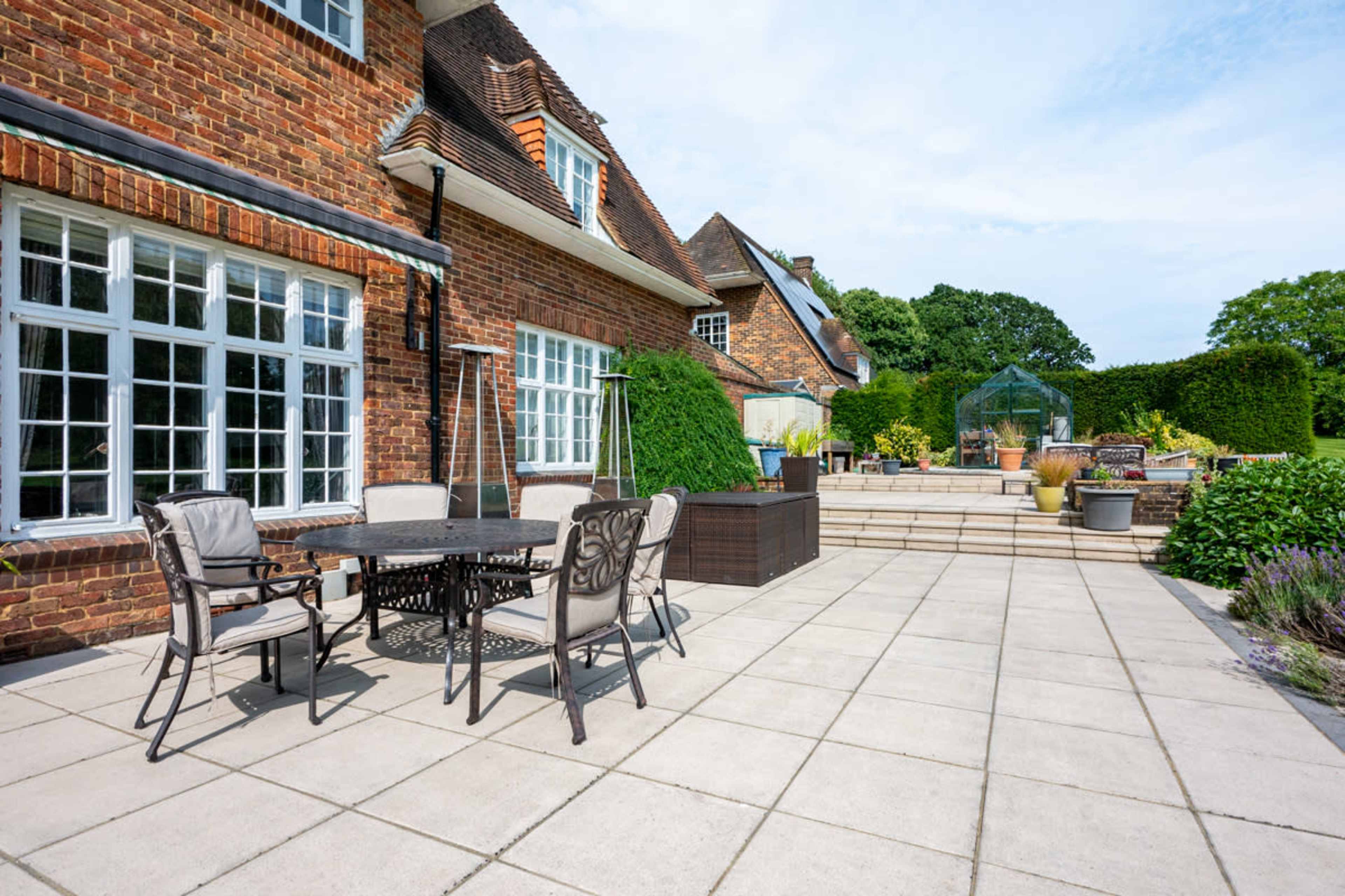 A brick house with a patio features a round table and chairs, surrounded by landscaped gardens and a greenhouse in the background.