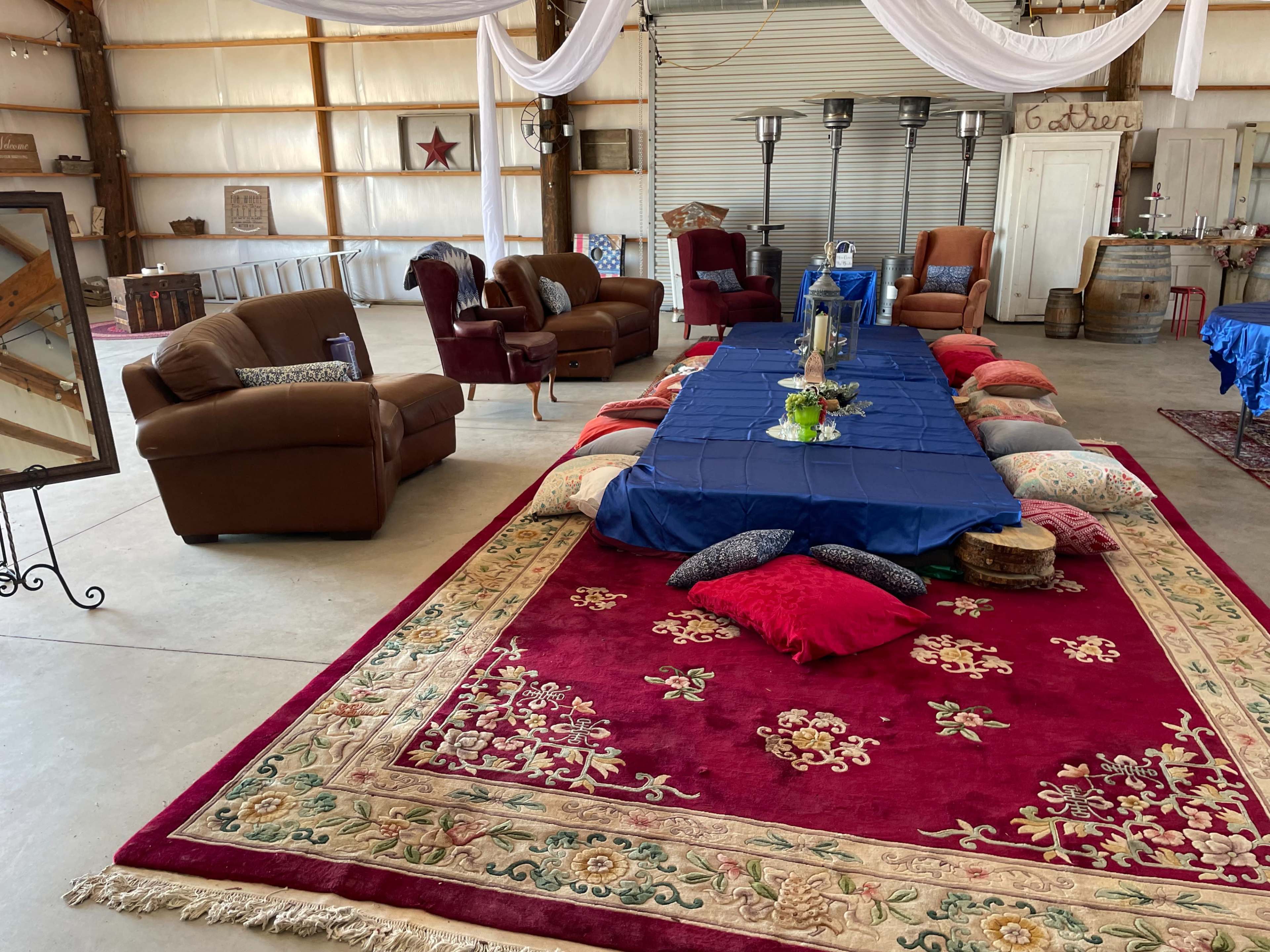 A spacious indoor setting with a long blue table surrounded by colorful cushions on a large patterned rug, flanked by seating areas with brown sofas.