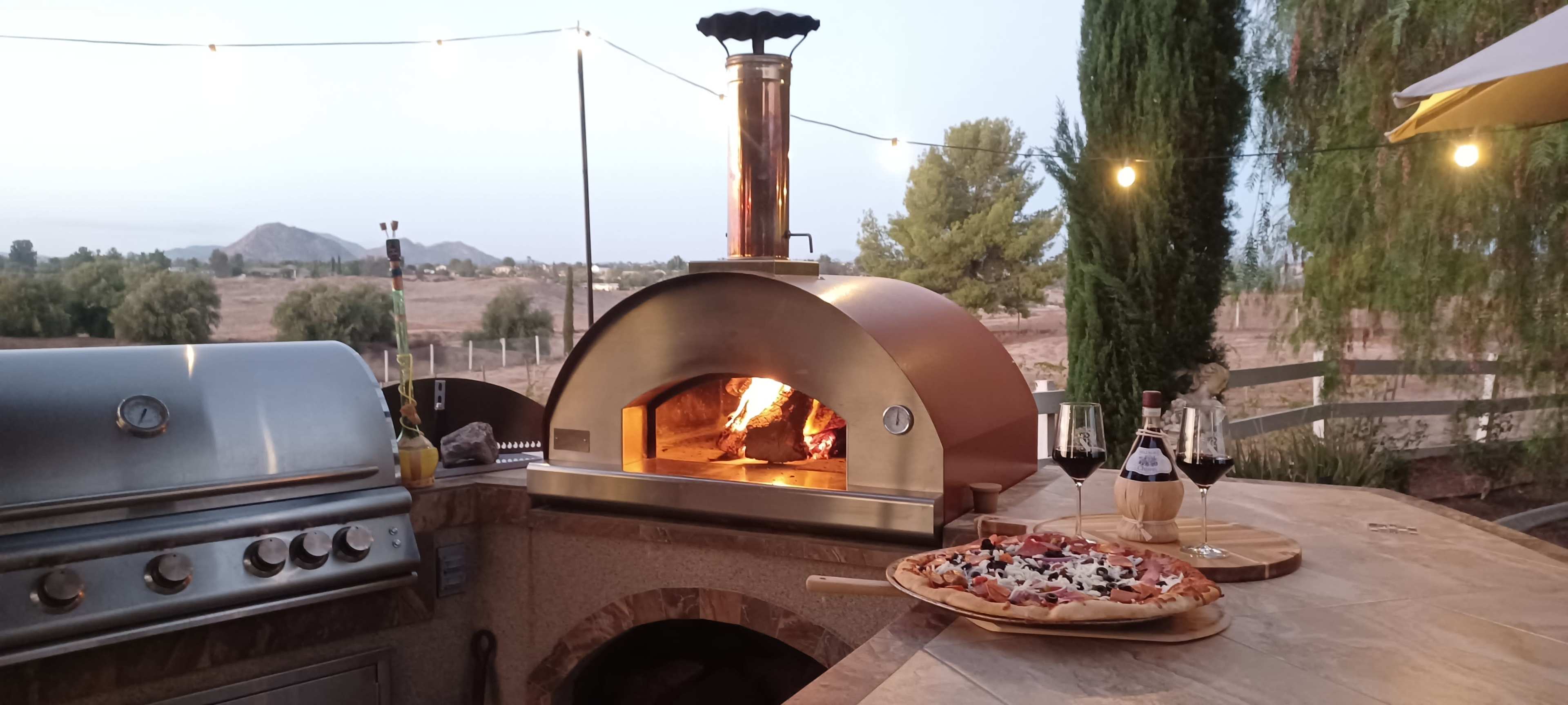 An outdoor kitchen features a stone pizza oven with flames visible inside, accompanied by a freshly baked pizza on the counter and two glasses of red wine nearby.